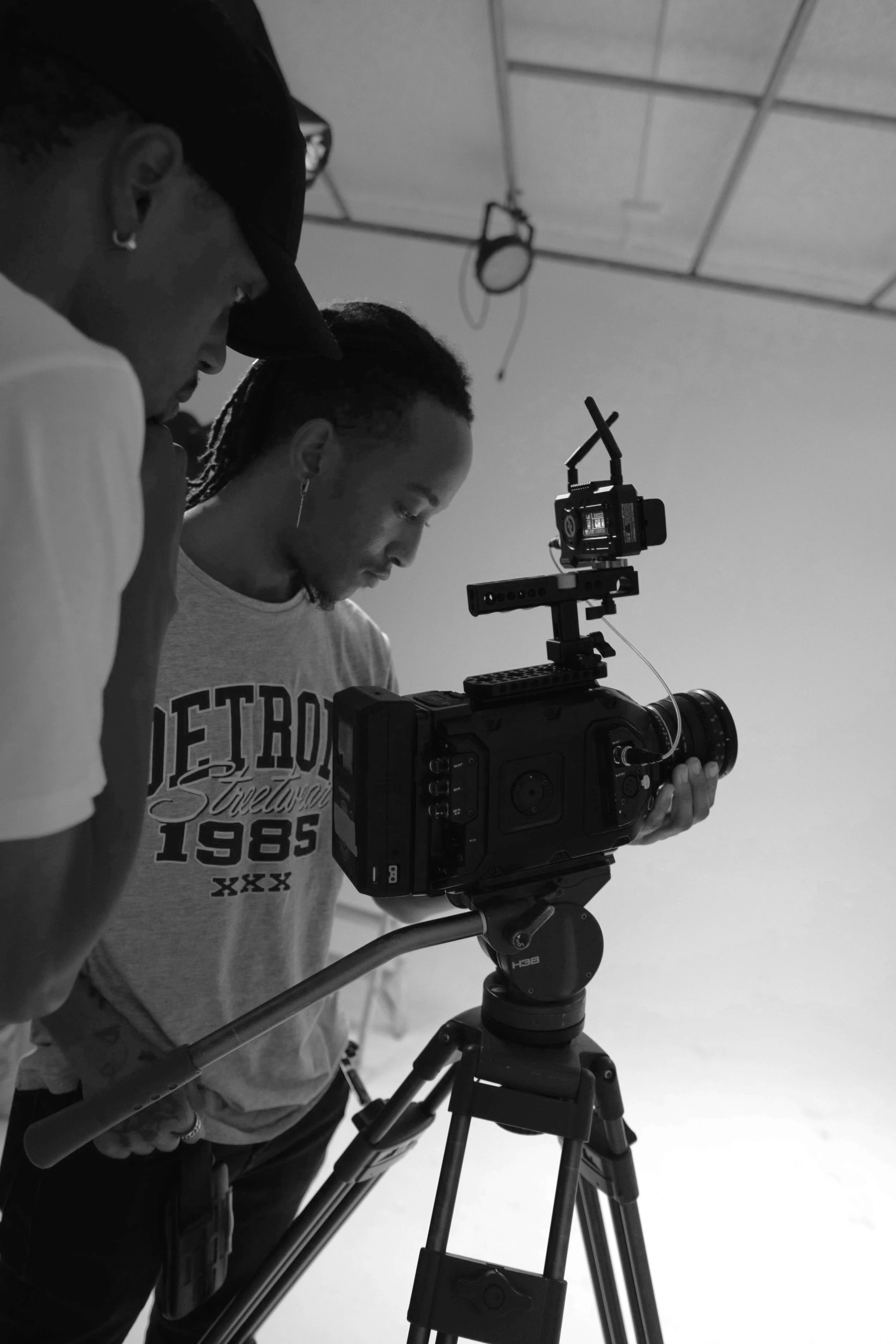 Two individuals operating a professional camera on a tripod in a studio with a plain background and ceiling lighting fixtures.