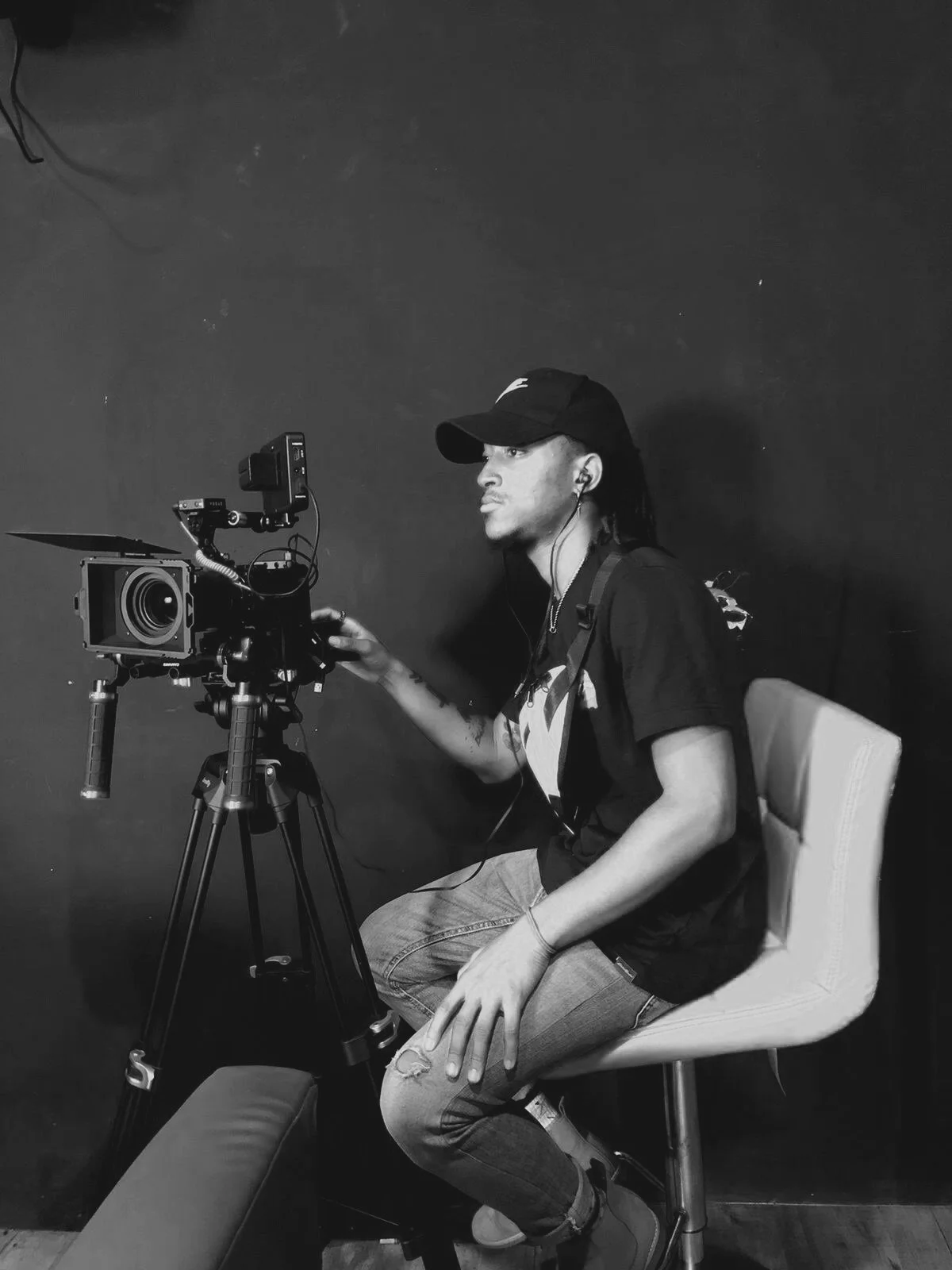 A young man sitting on a chair in front of a camera, wearing a cap, t-shirt, ripped jeans, and earphones, in a black-and-white photo.