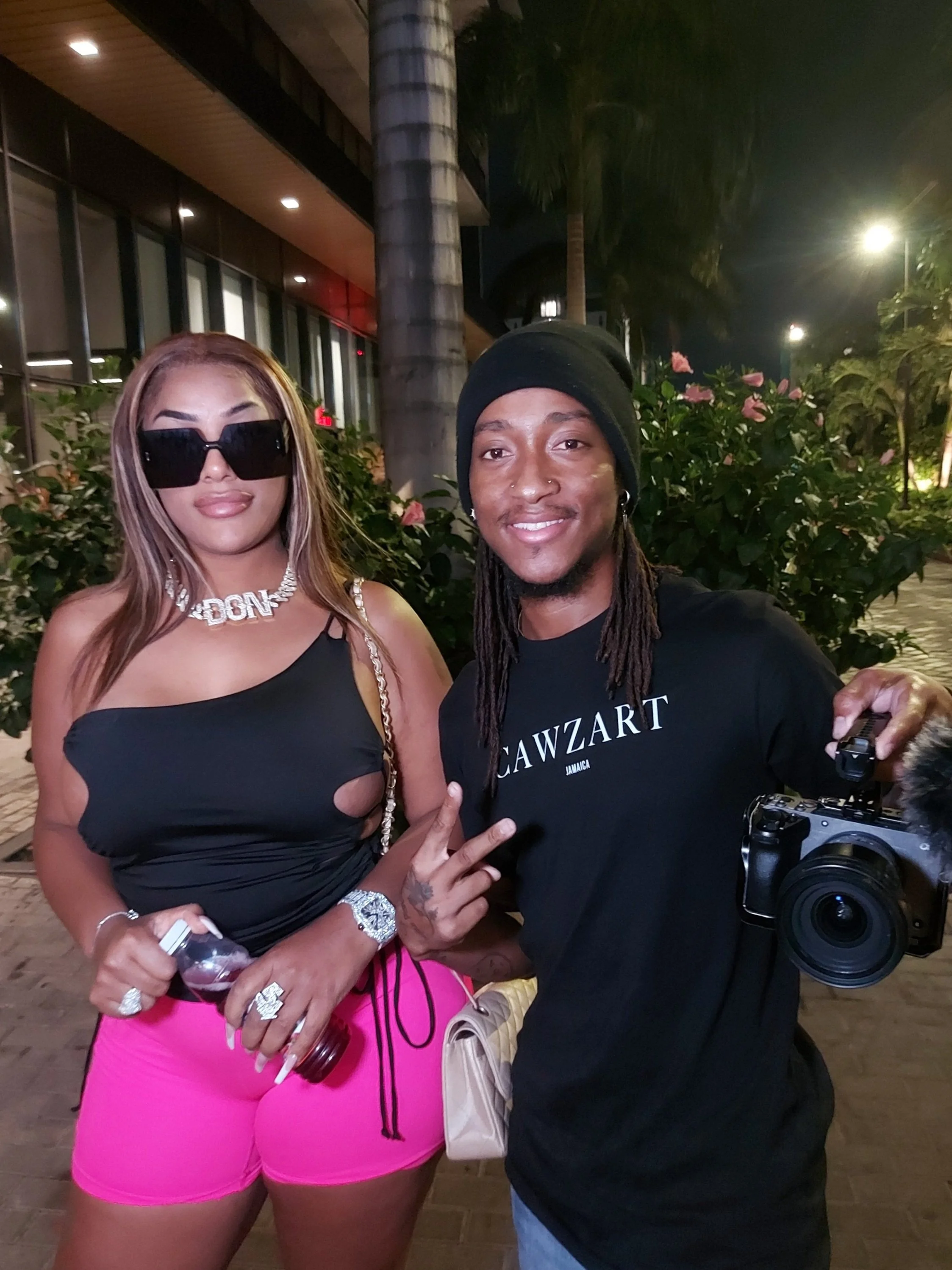 Two people posing for a photo outdoors at night. The woman on the left is wearing large black sunglasses, a black top, pink shorts, and jewelry, holding a bottle of wine and a cell phone. The man on the right is wearing a black beanie, a black T-shir