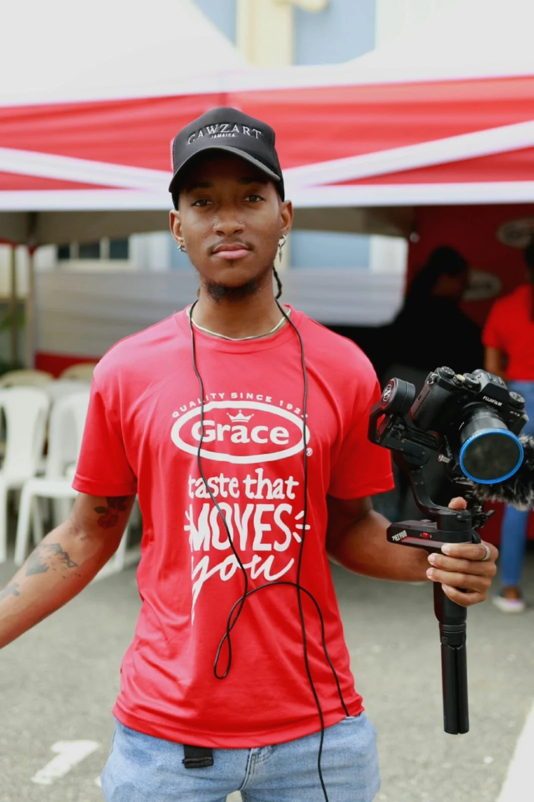 A young man holding a camera, wearing a red t-shirt with graphic text, a black cap, and earphones, standing outdoors under a red and white canopy.