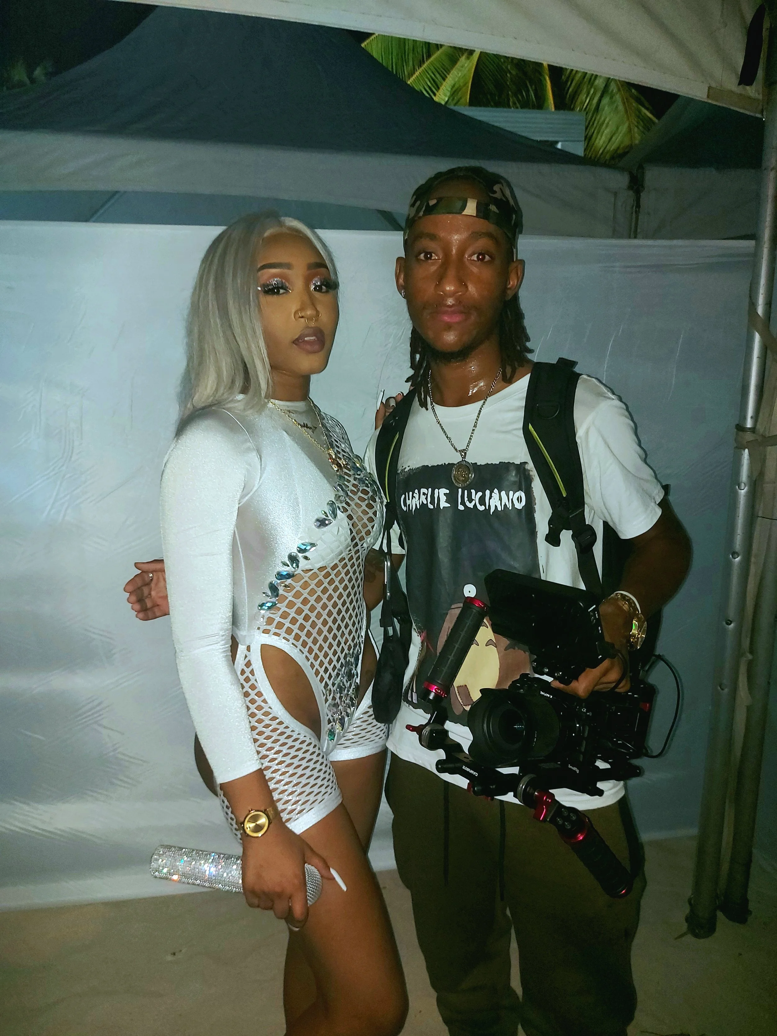 A woman with platinum blonde hair, wearing glamorous makeup, a white outfit with cut-out details, and holding a sparkly clutch, poses next to a man with dreadlocks, wearing a white t-shirt with Charlie Luciano written on it, a patterned cap, and hold