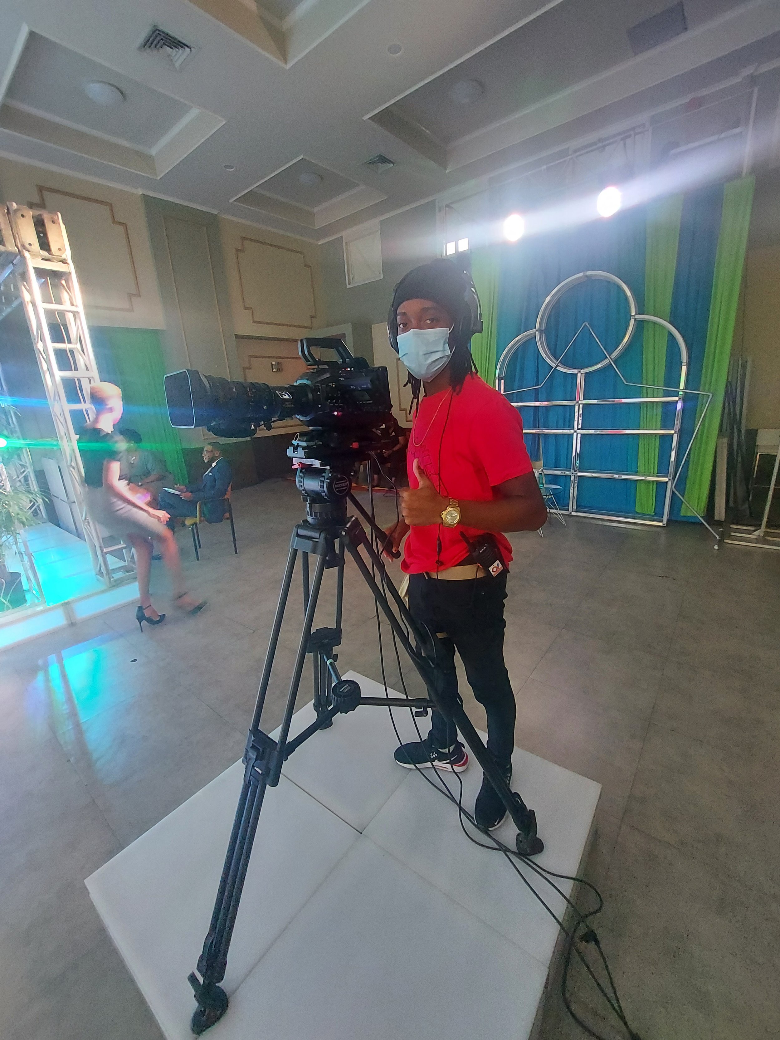 A person wearing a black beanie hat, face mask, red T-shirt, and headphones standing behind a professional video camera on a tripod, capturing a shot at an indoor event with colorful blue and green curtains and stage lighting.