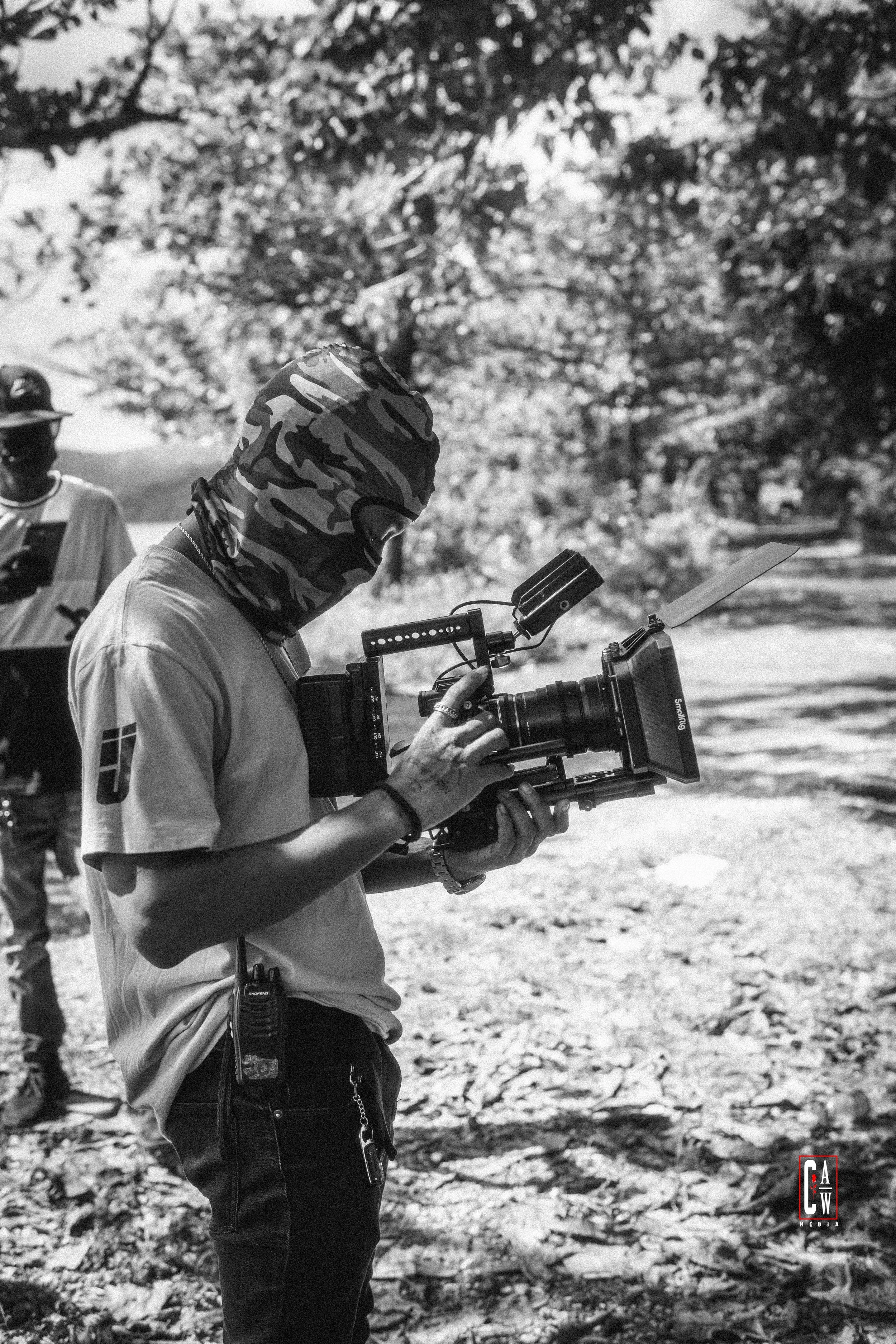 A person with a camouflage face covering and wearing a cap is filming or taking photos with a professional camera outdoors. Two other individuals are visible in the background, one on the left wearing glasses and a cap, and the other partially visibl