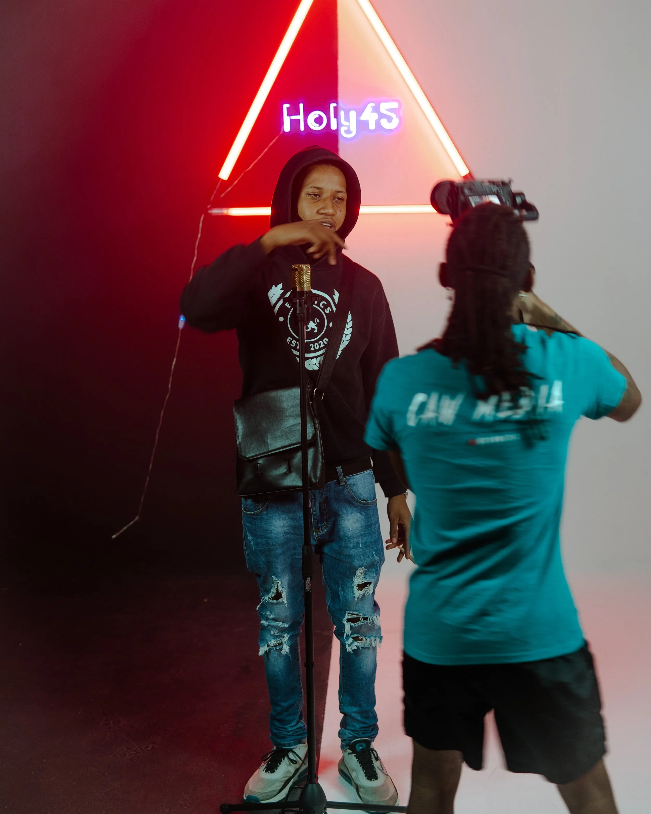 A young man with a hoodie and ripped jeans standing in front of a neon triangle sign reading 'Holy45', being filmed by a person with braided hair, wearing a turquoise shirt, in an indoor setting with the left side of the background dark and the right