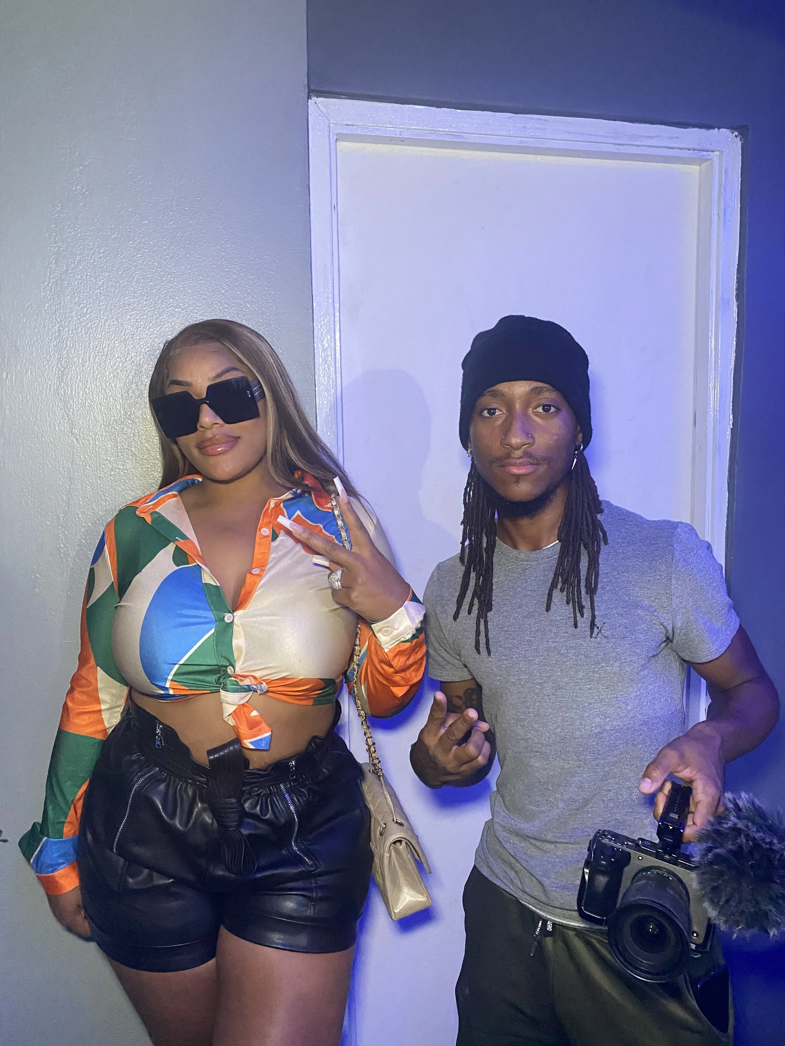 A woman wearing large black sunglasses, a colorful patterned shirt tied at the waist, and black leather shorts, standing next to a man with dreadlocks, a black beanie, gray T-shirt, and holding a camera. Both are posing with relaxed expressions again