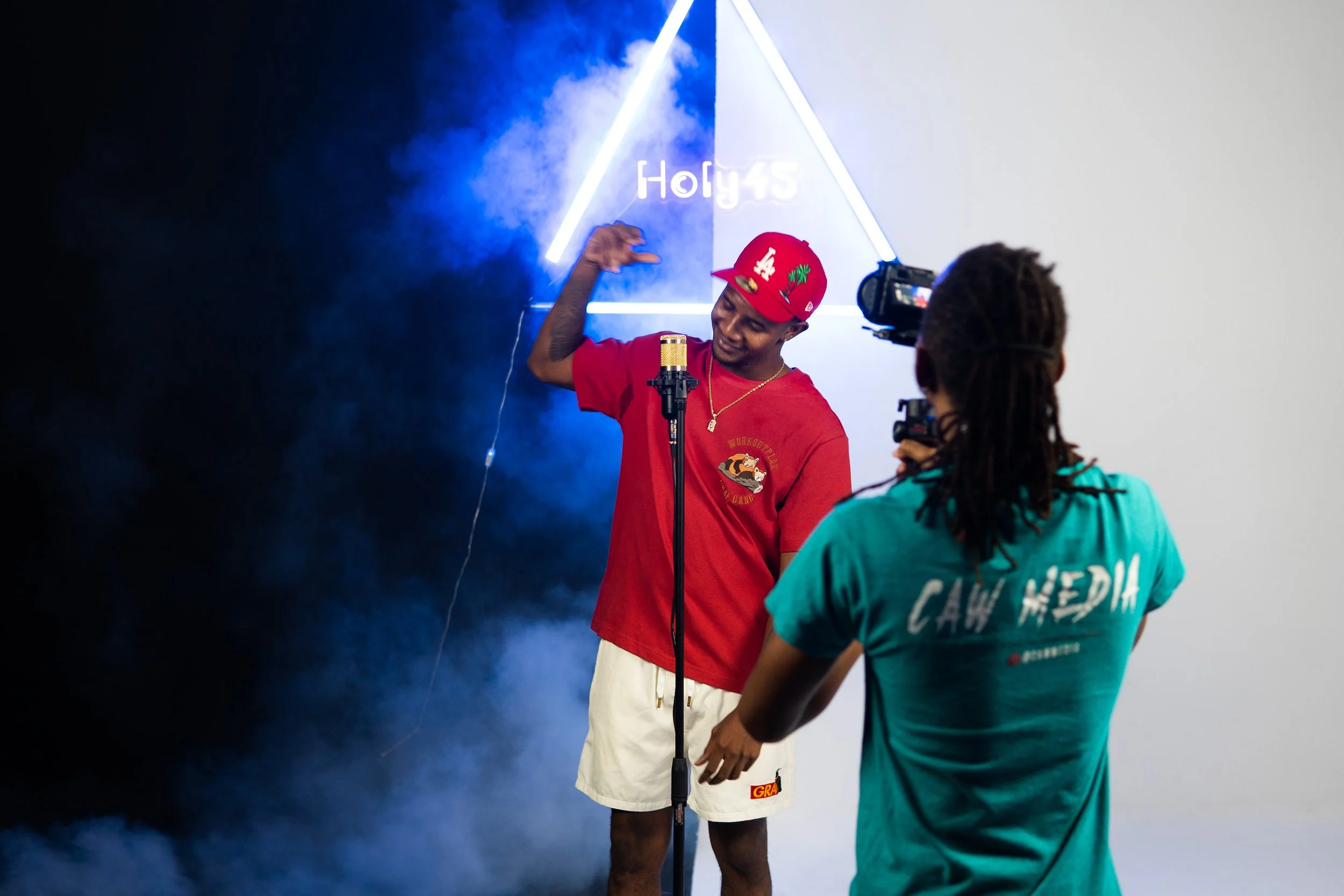A man wearing a red cap and red t-shirt is posing with a smile in front of a blue and white background with neon lights. A person with braided hair is filming him with a camera.