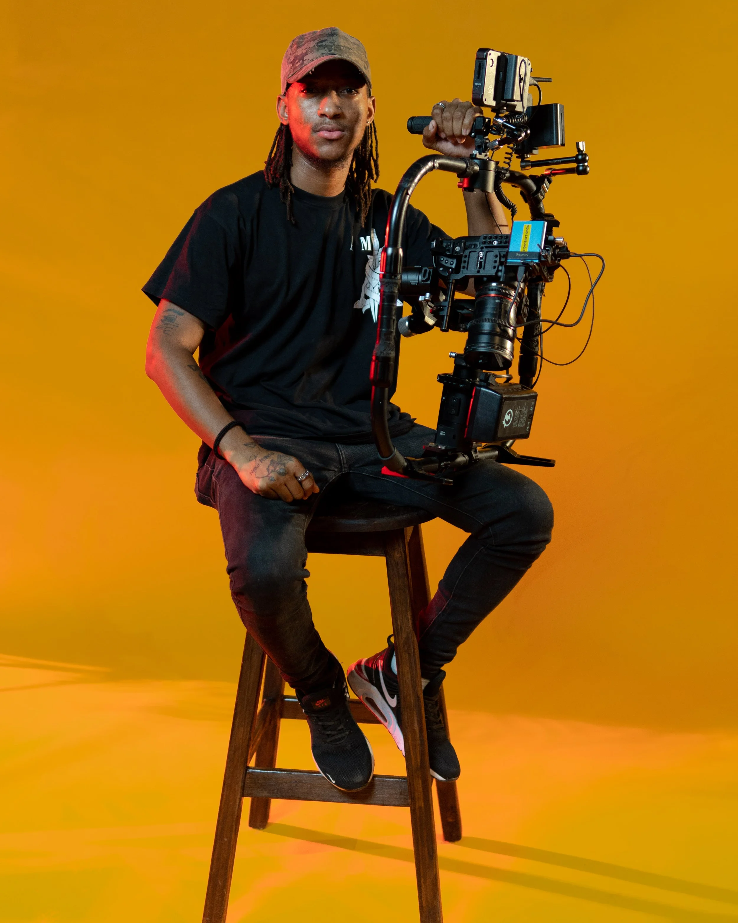 A young man with dreadlocks wearing a camouflage cap, black t-shirt, and black jeans, sitting on a wooden stool holding a camera stabilizer setup against a yellow background.