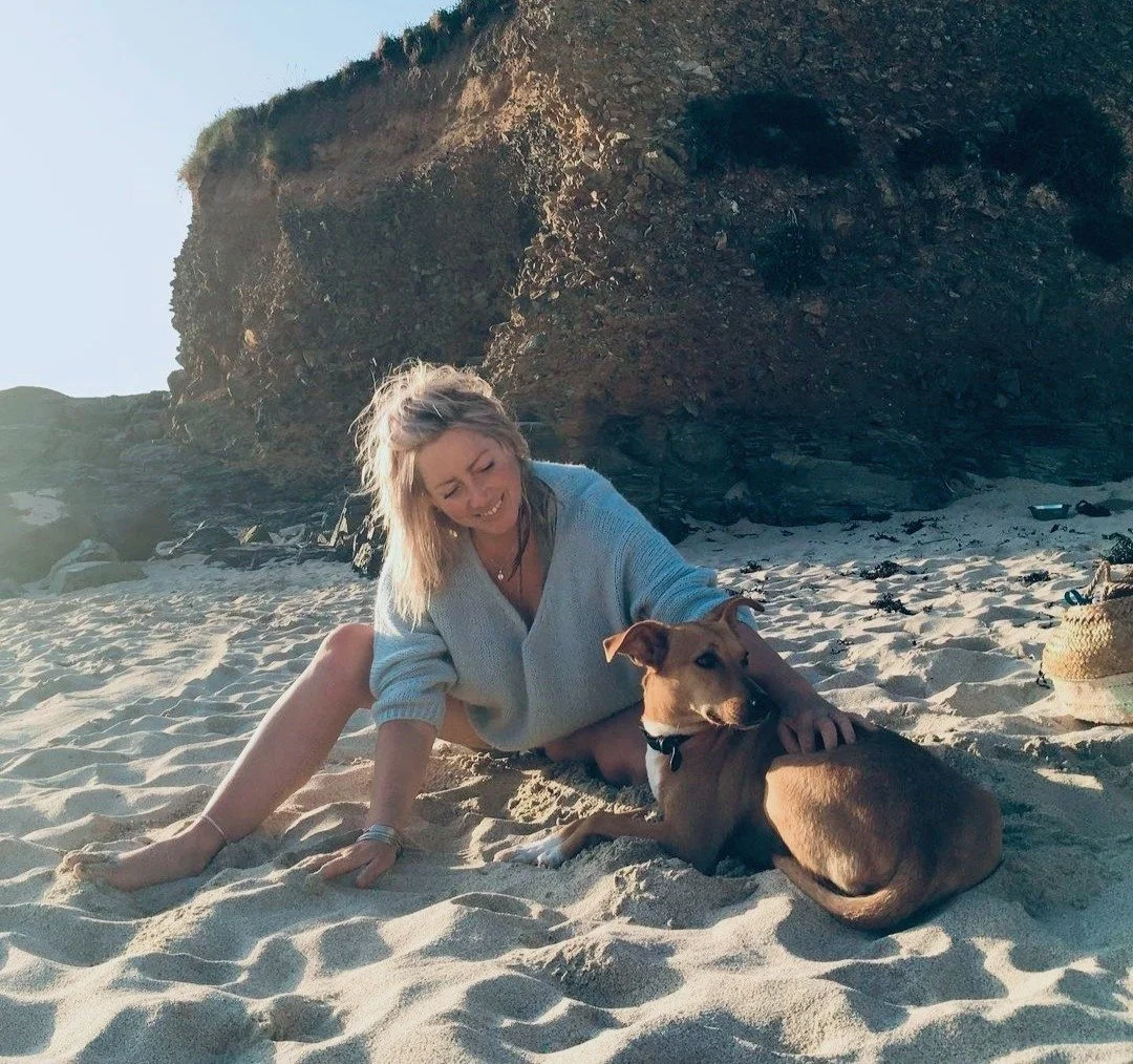 A woman with blonde hair in a light-colored sweater sitting on sandy beach next to a brown dog, with large rock formations in the background.