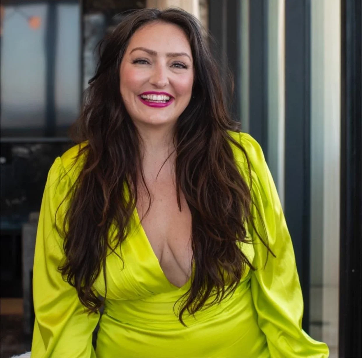 A woman with long wavy brown hair smiling, wearing a bright yellow dress with a deep V-neck, standing in front of a modern building with glass windows.