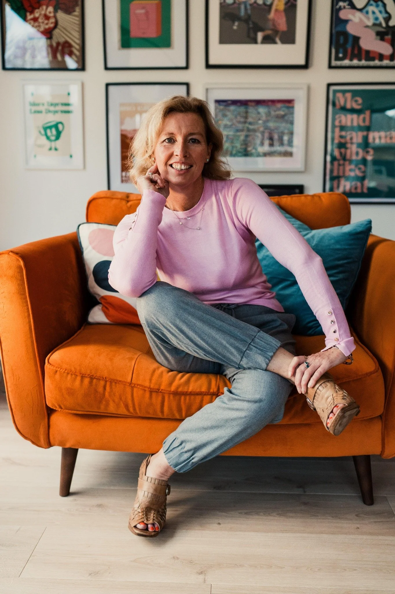 A middle-aged woman with blonde hair sitting comfortably on an orange couch in a colorful, art-filled room, smiling at the camera.