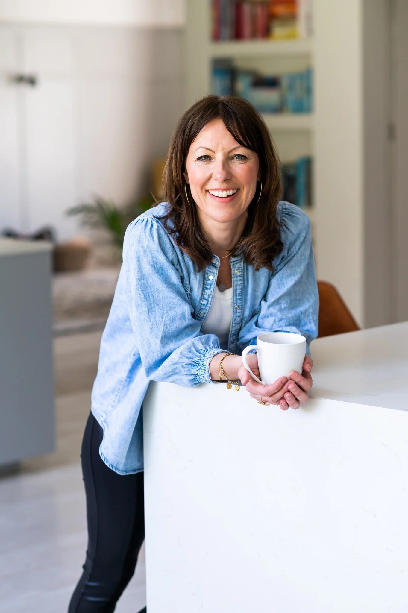 A woman with brown hair smiling and holding a white mug in a modern kitchen.