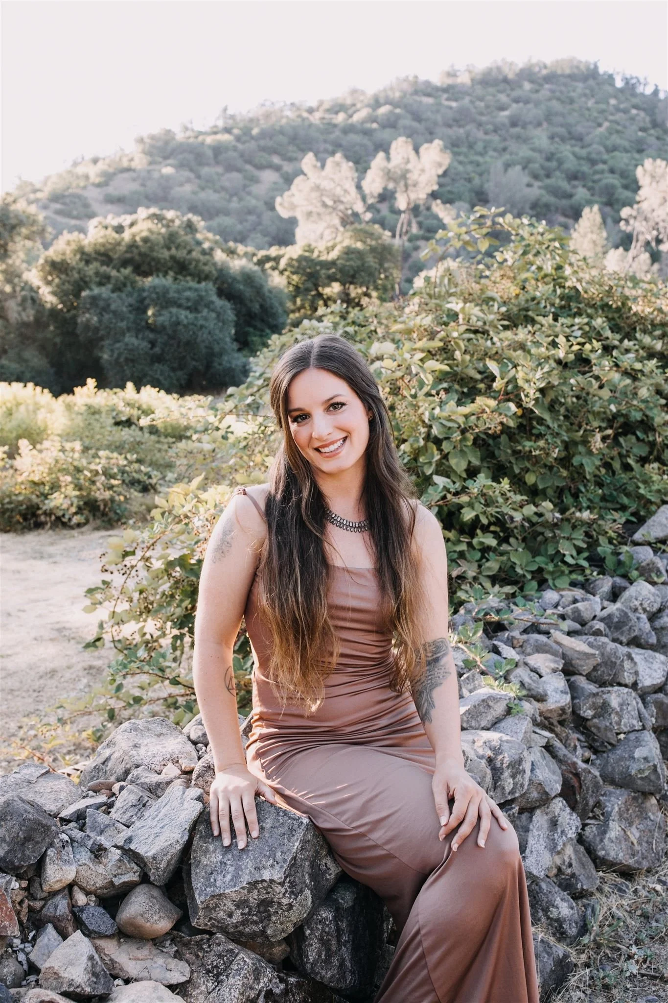 Young woman with long brown hair, wearing a brown satin dress and a necklace, sitting on rocks outdoors with greenery and hills in the background, smiling at the camera.