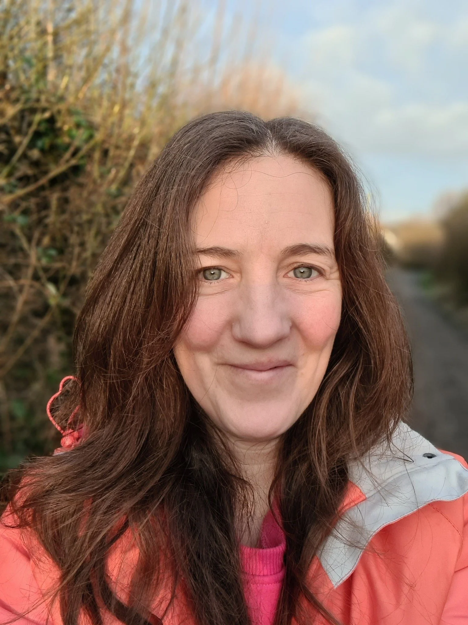 A woman with long brown hair, light skin, and green eyes standing outdoors on a path with sunlight. She is wearing a coral jacket and pink top, smiling at the camera.