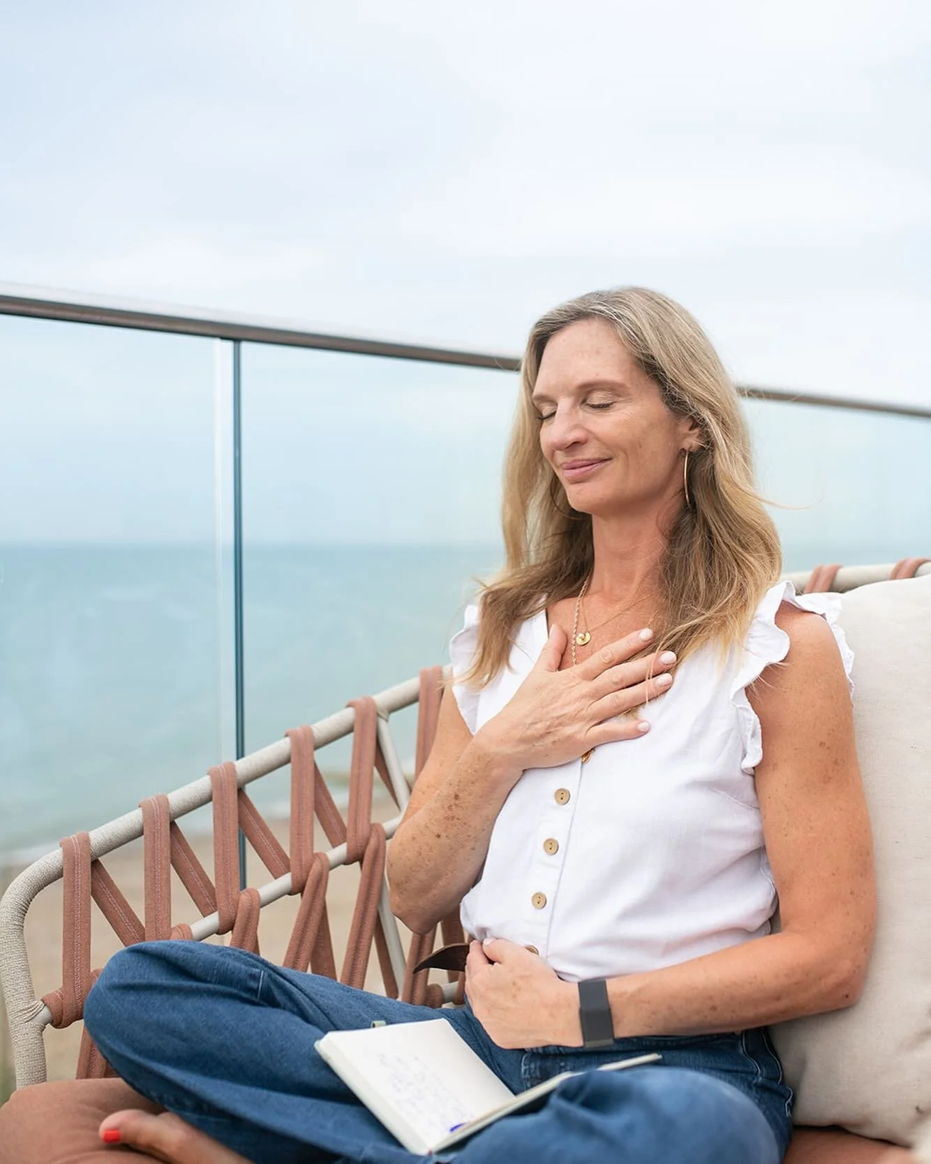 A woman sitting on a balcony with her eyes closed, smiling, holding her hand over her chest. She has long blonde hair, wears a white sleeveless blouse, blue jeans, and a black wristband. There is a notebook on her lap, and the background shows a view of the ocean and a cloudy sky.