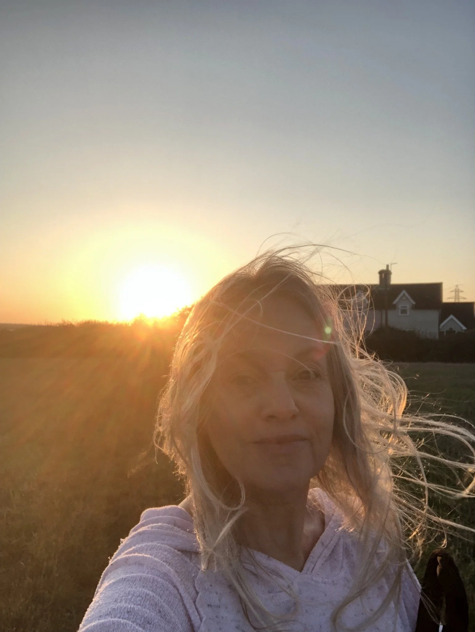 A woman with blonde hair taking a selfie outdoors during sunset with a house and power lines in the background.