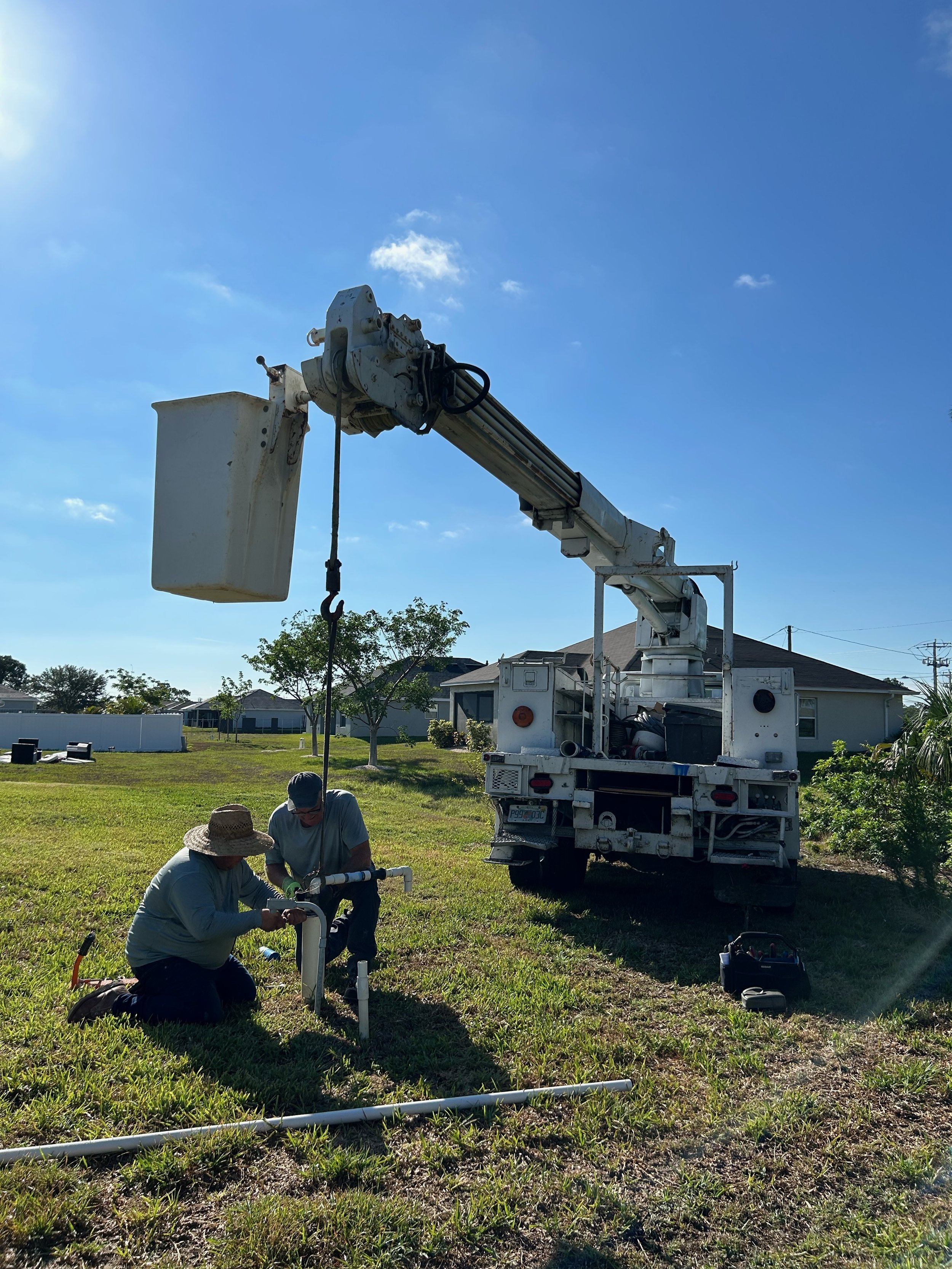 Two workers installing or repairing an outdoor water pipe connection near a utility truck in a grassy yard on a sunny day.