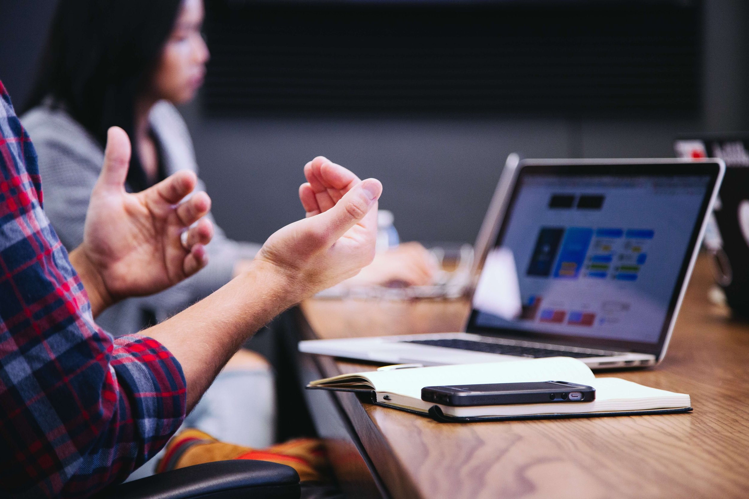 A close up on someone's hands as they express themselves in a meeting. A laptop is close to their hands along with a notebook which has a mobile phone placed on top.