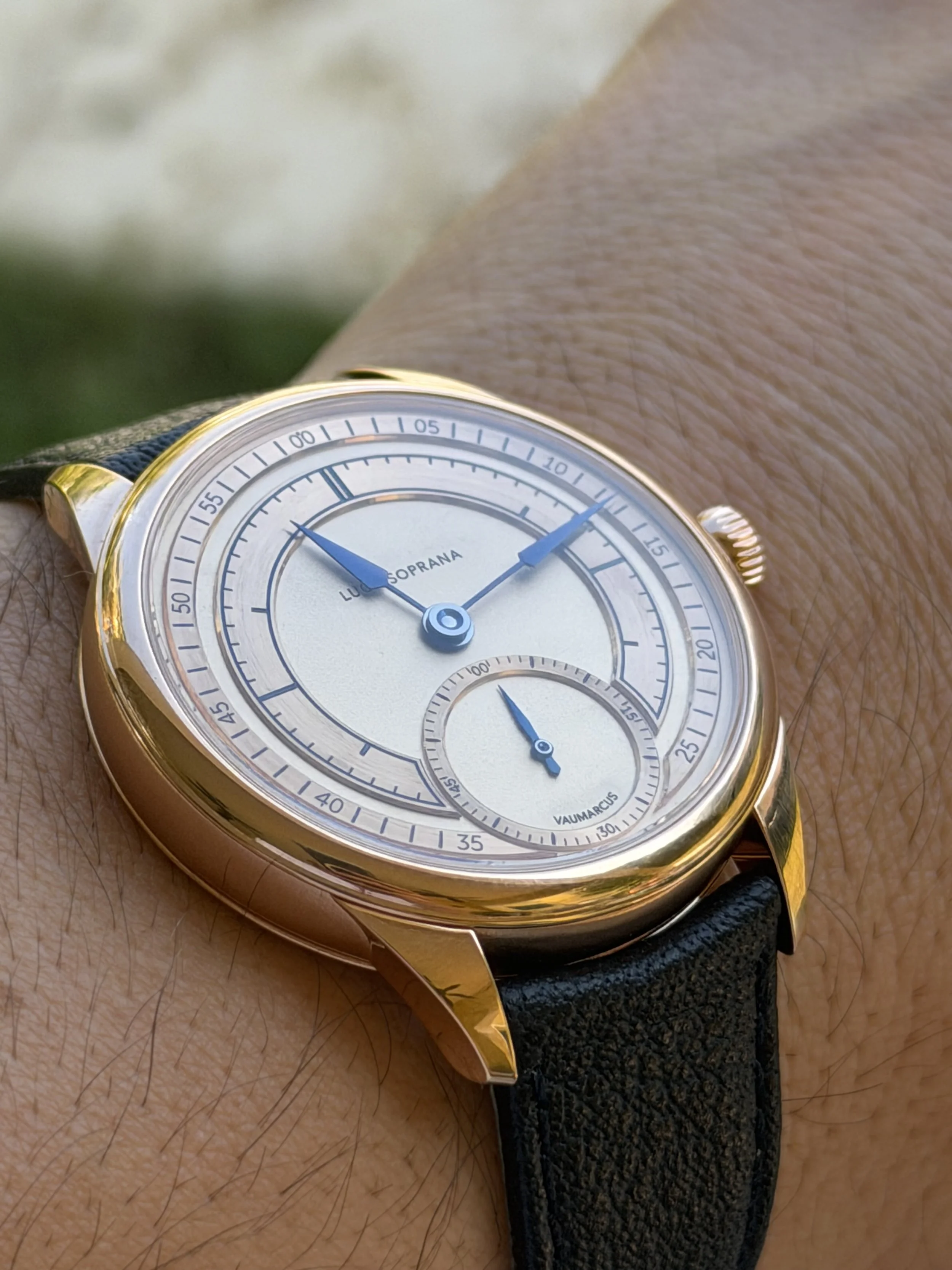 Close-up of a luxury wristwatch on a person's wrist, featuring a white face, blue hands, and a black leather strap.