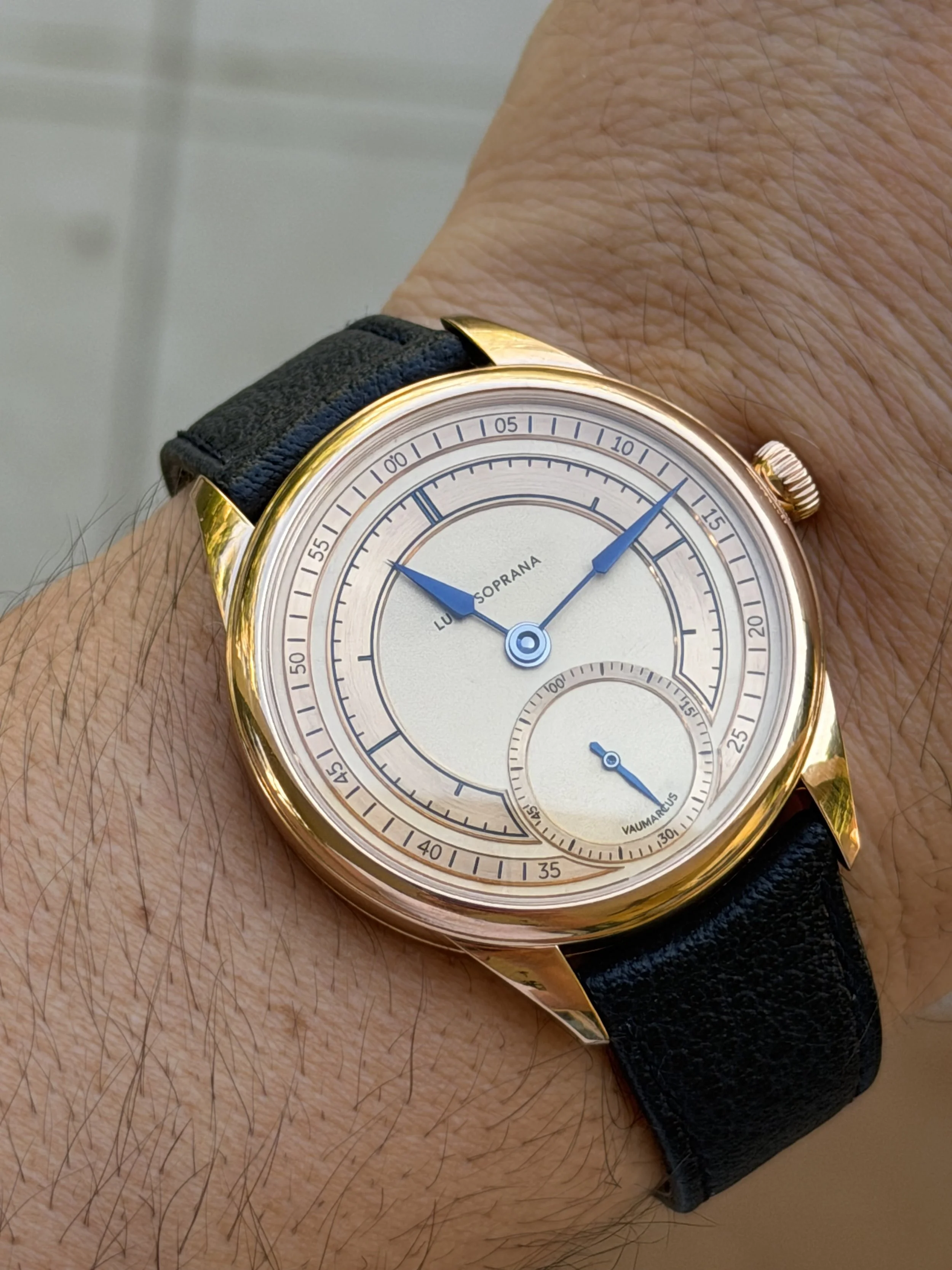 Close-up of a wristwatch with a gold case, black leather strap, and a cream dial featuring blue hands. The watch has multiple subdials and markings, with the brand name "LUCIEN SOPRANA" visible on the main dial.
