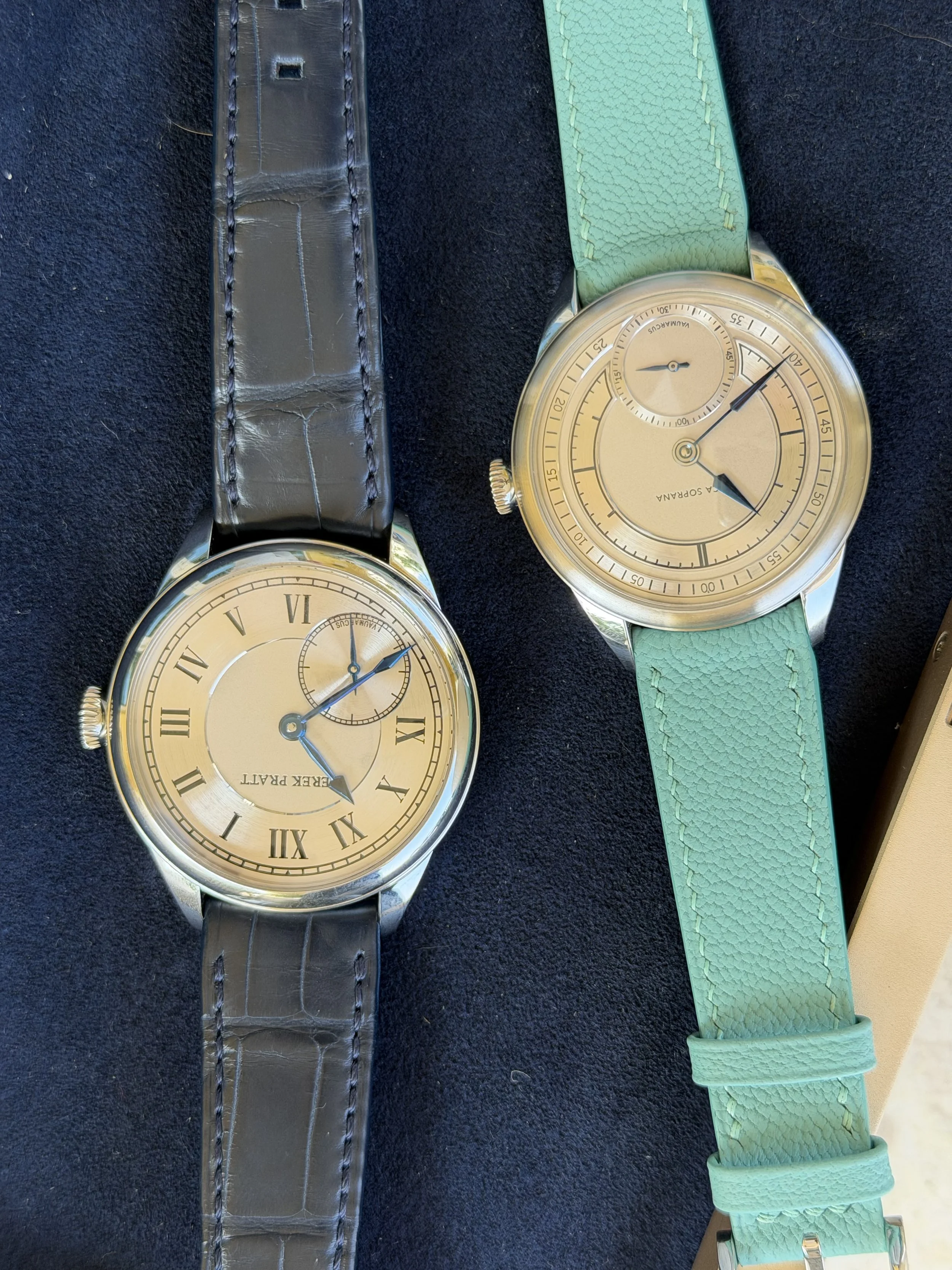 Two wristwatches with leather straps on a black surface. The watch on the left has a black strap and a beige dial with Roman numerals. The watch on the right has a green strap and a beige dial with a small sub-dial and a power reserve indicator.