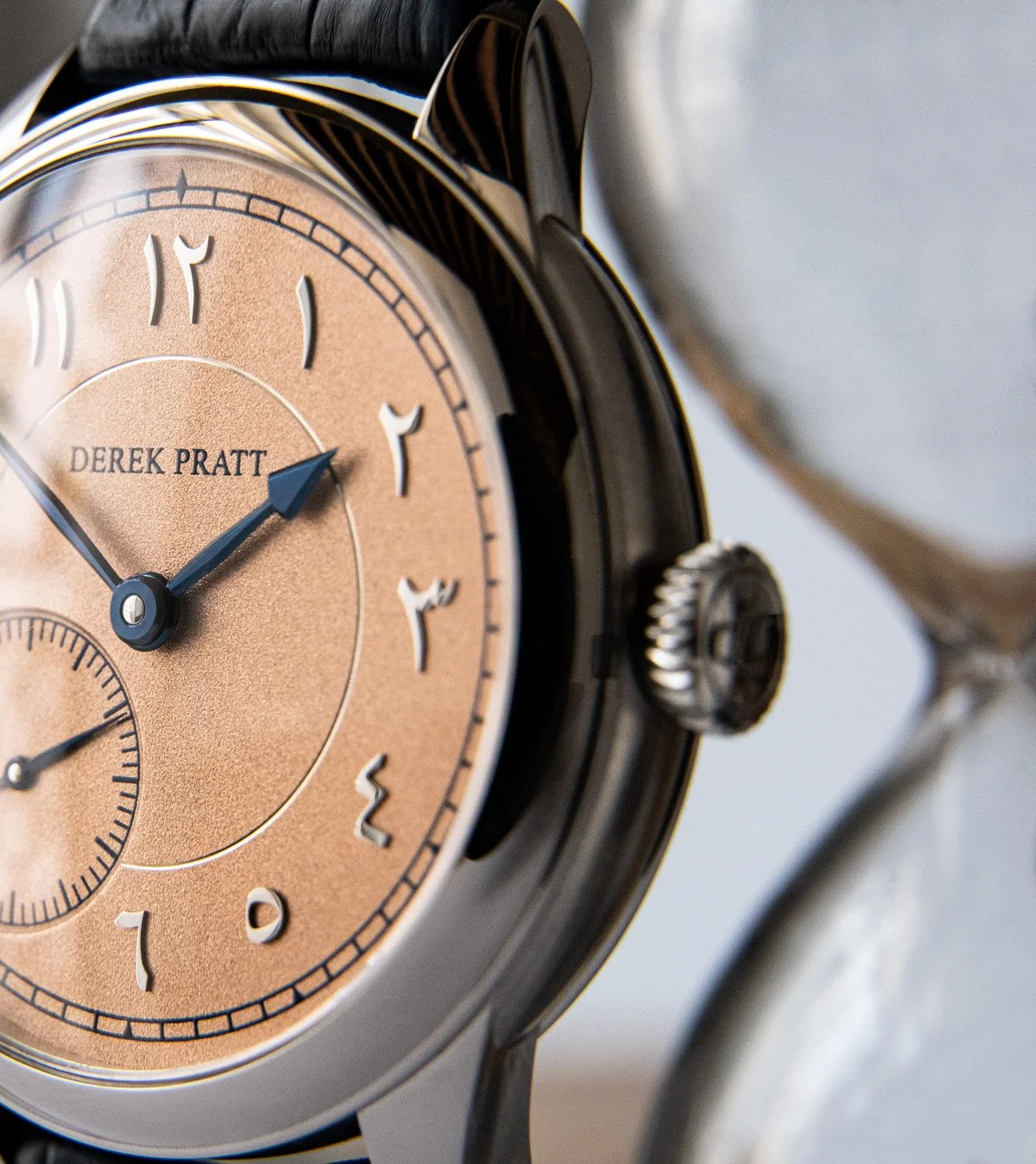 Close-up of a luxury wristwatch with a beige dial, black hour and minute hands, and a small seconds subdial, with the name Derek Pratt on the face.