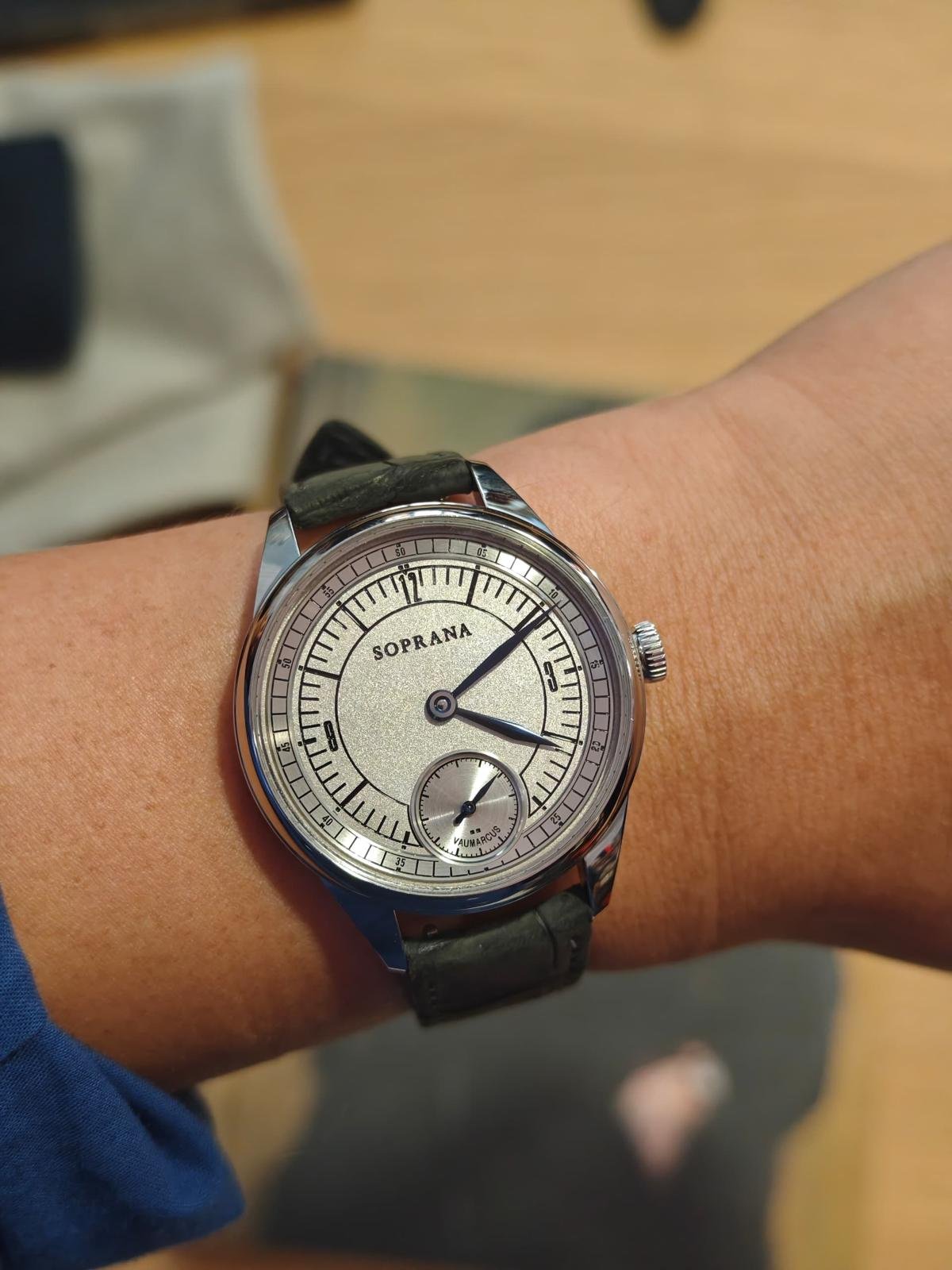 A wristwatch with a silver dial, black leather strap, and the word 'SOPRANA' in the center. The watch shows the time as 2:09 with a small subdial at the 6 o'clock position.