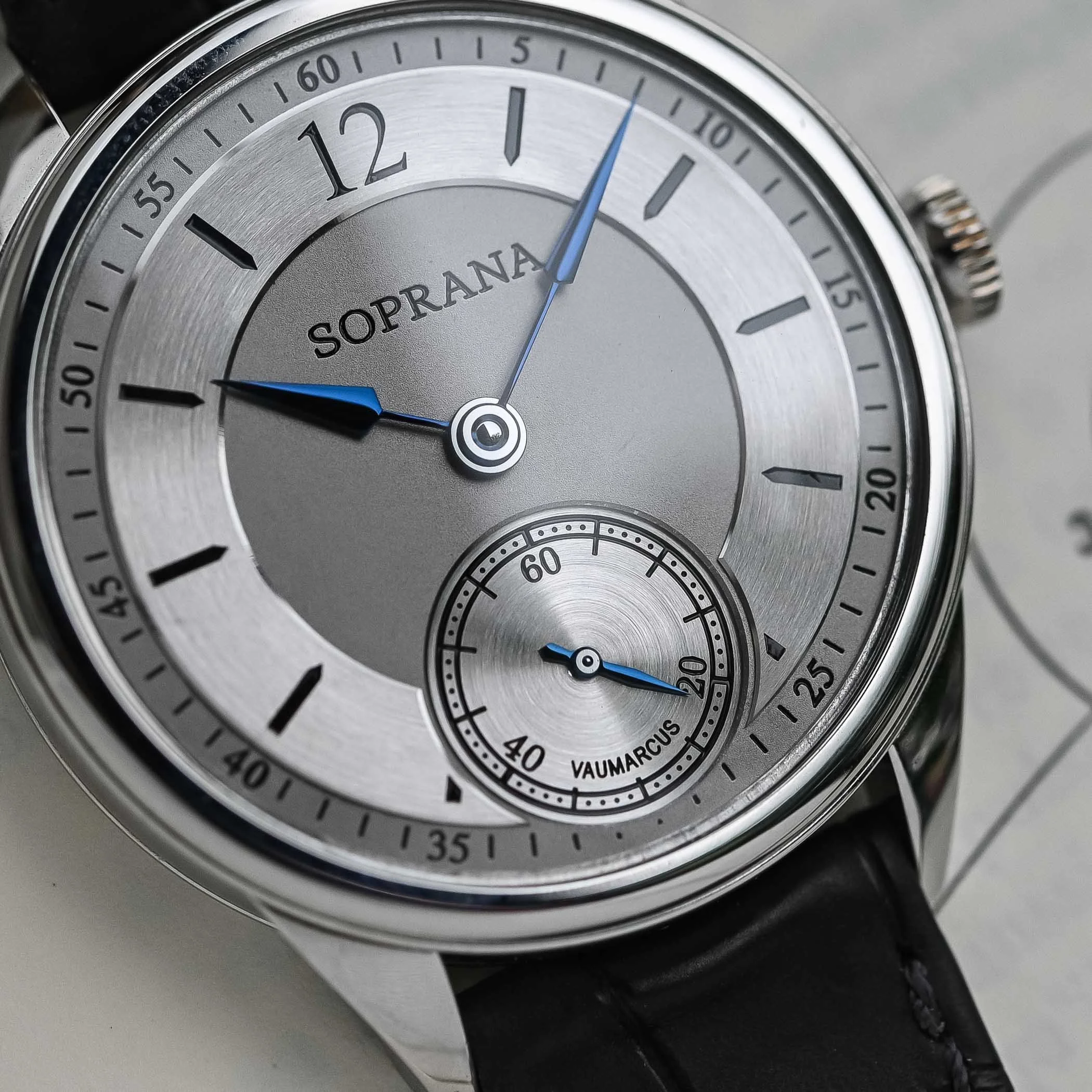 Close-up of a silver wristwatch with the brand name SOPRANA on the dial, featuring blue hour and minute hands, a small seconds subdial at the 6 o'clock position, and a black leather strap.