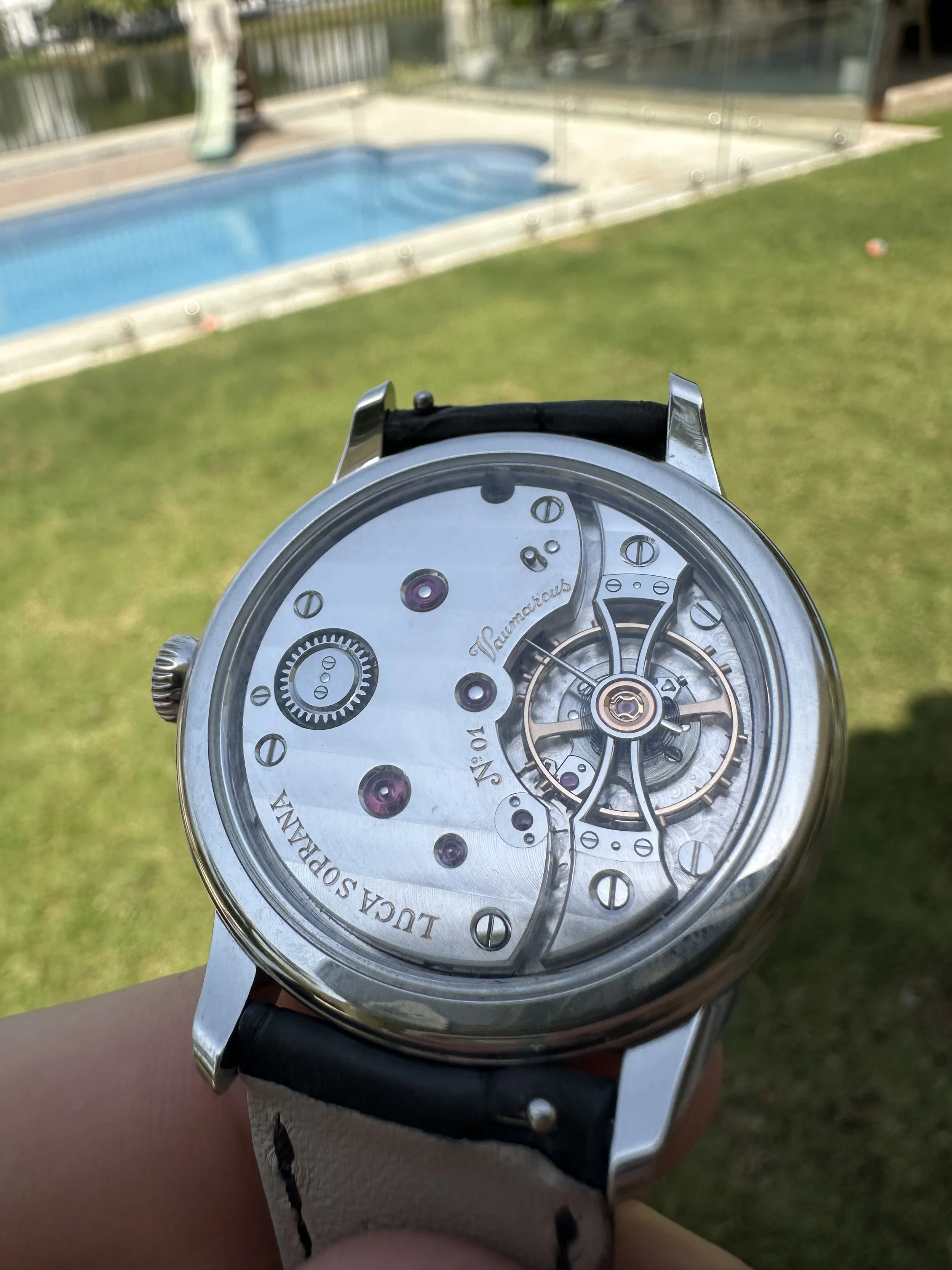 Close-up of a mechanical wristwatch with an exposed inner mechanism, held outdoors near a grassy area with a swimming pool in the background.