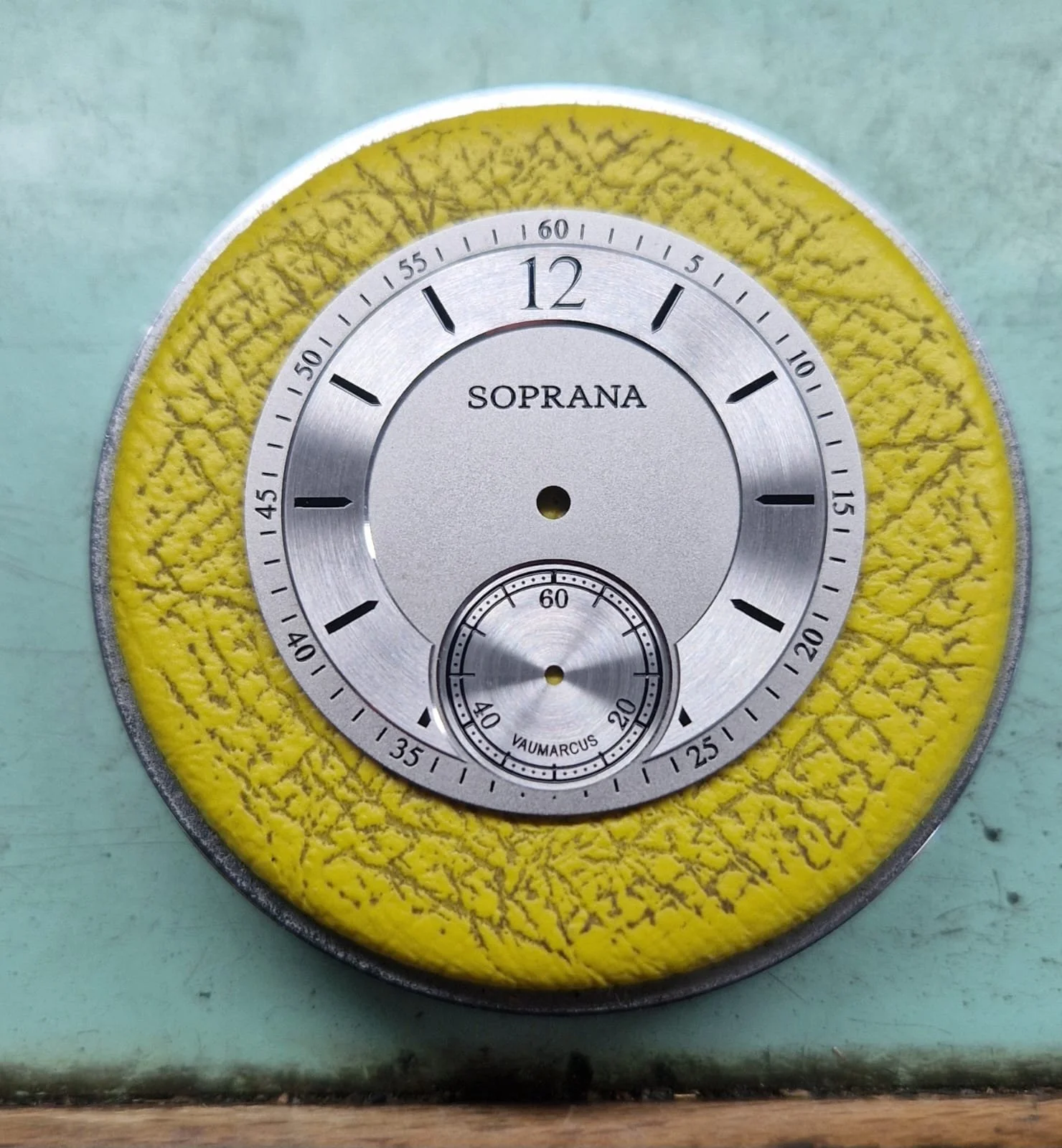 Close-up of a vintage Soprana watch face with a yellow textured background, showing the time and a small seconds dial.