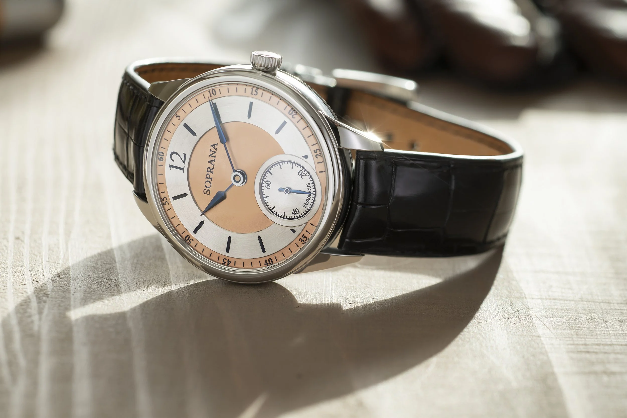 A luxury wristwatch with a black leather strap, silver case, and a beige and silver dial with sub-dials, on a light wooden surface.