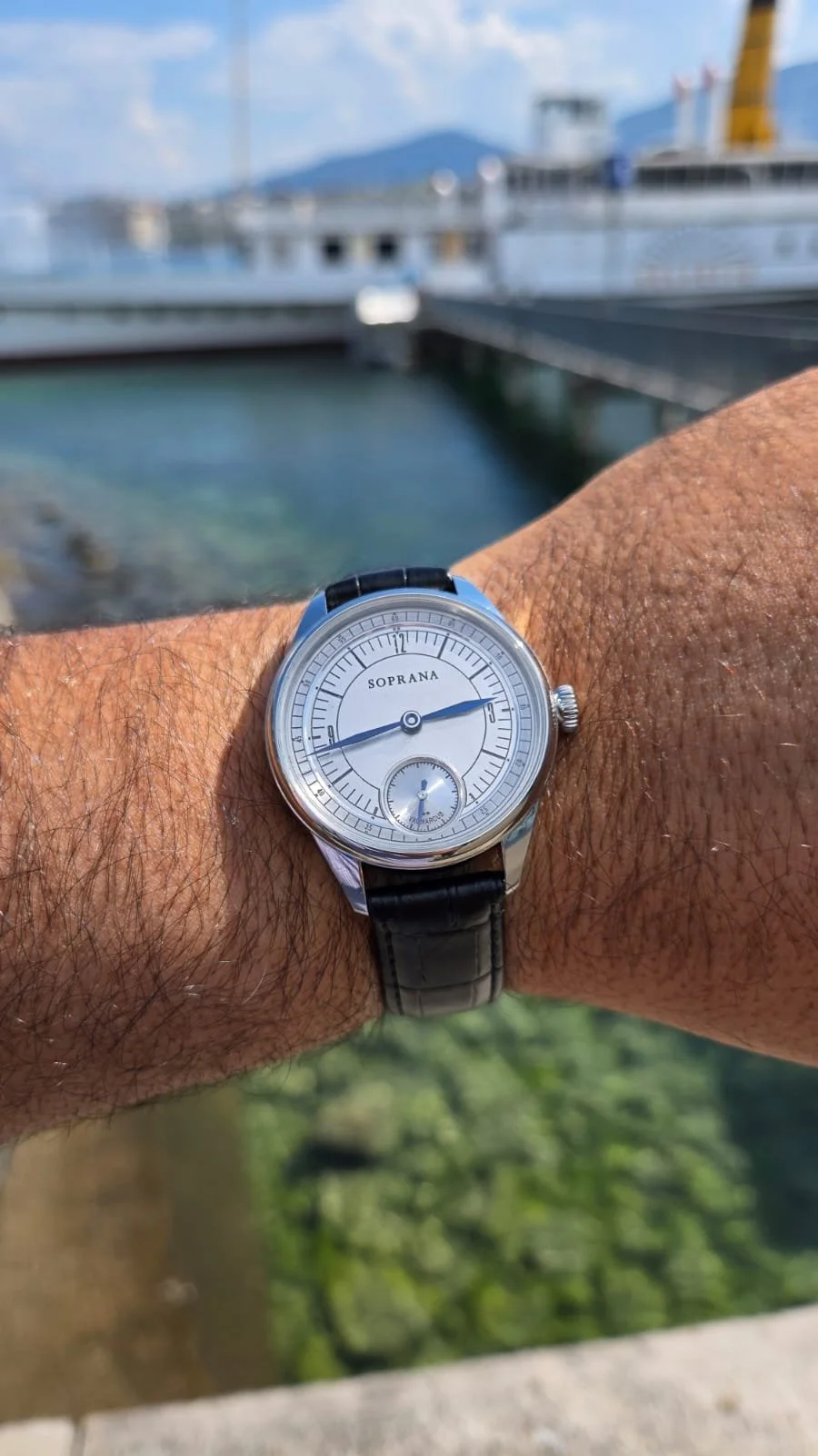A wristwatch with the brand name SOPRANA on a person's wrist. The watch has a silver case, white dial, and black leather strap. The time shown is approximately 3:52, with a smaller subdial at the 6 o'clock position. In the background, there is a mari