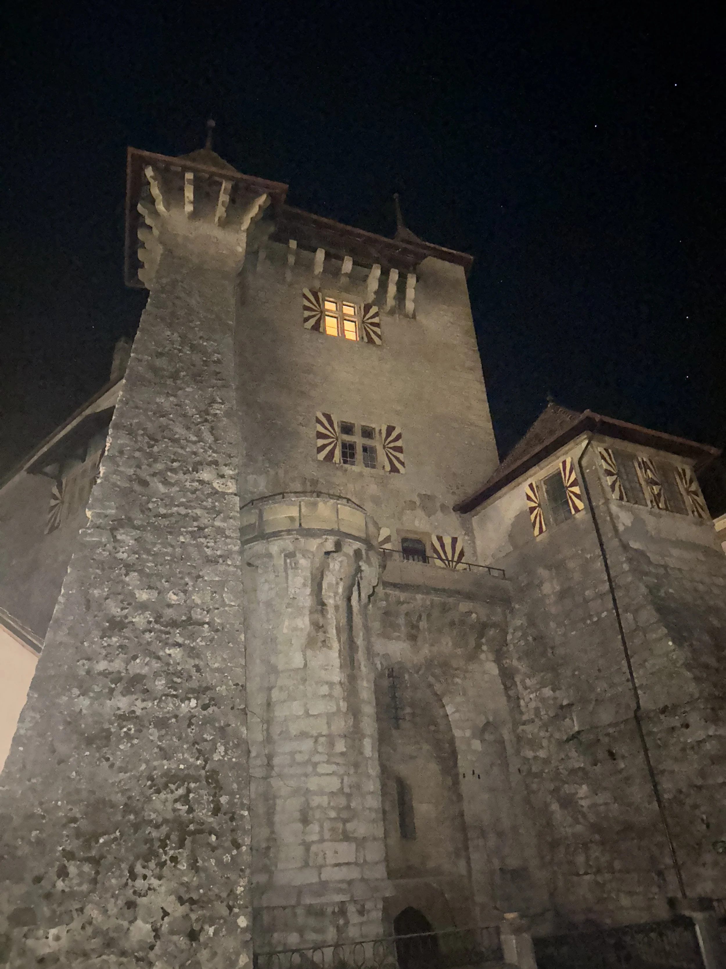 Historic stone castle tower at night with illuminated windows and decorative shutters, cloudy sky, and stars visible.