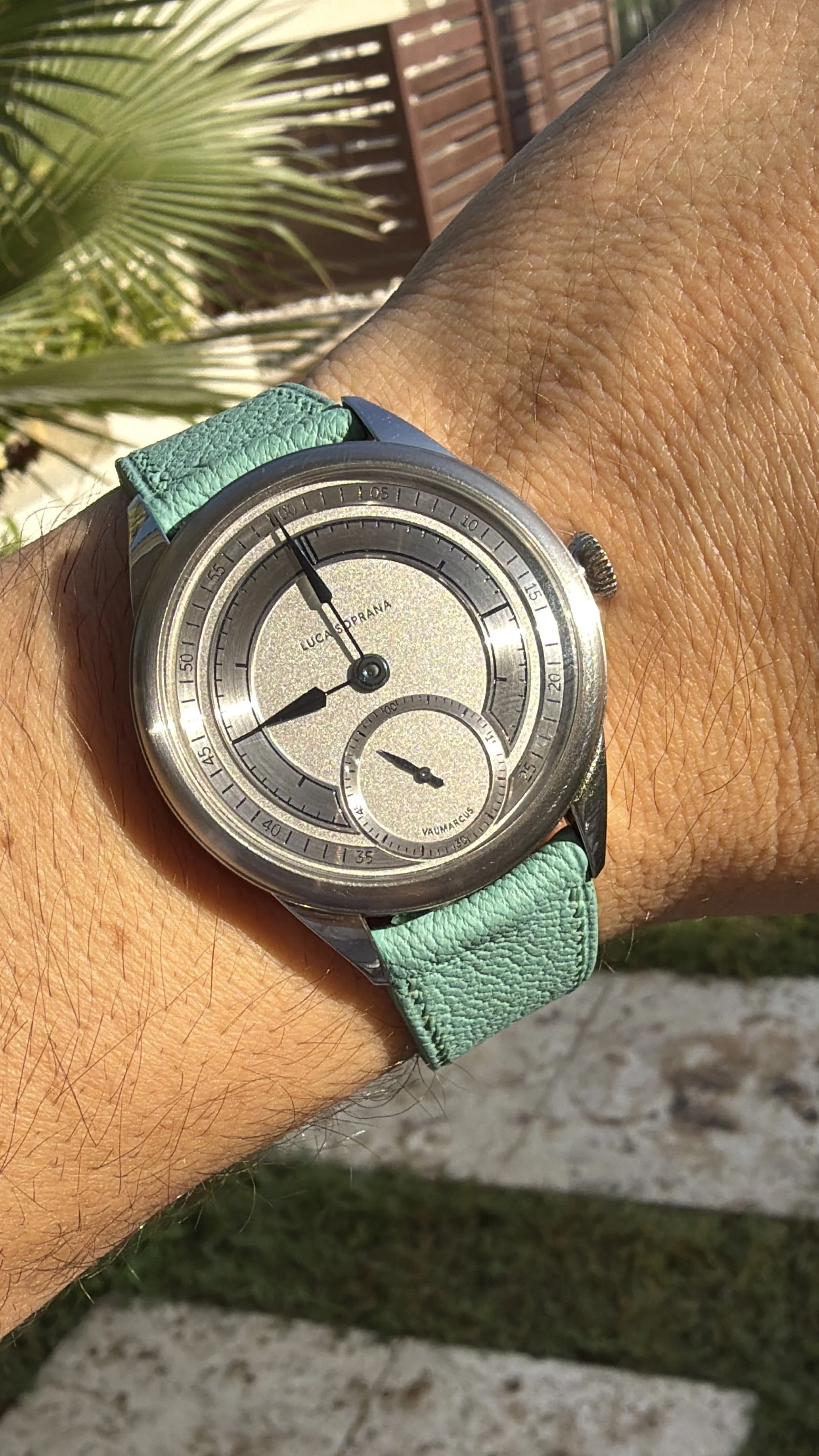 A wristwatch with a round silver face and black case, featuring two black hands for hours and minutes, and a small sub-dial with a black hand. The watch has a light green textured strap. The background includes a palm frond and a wooden fence.
