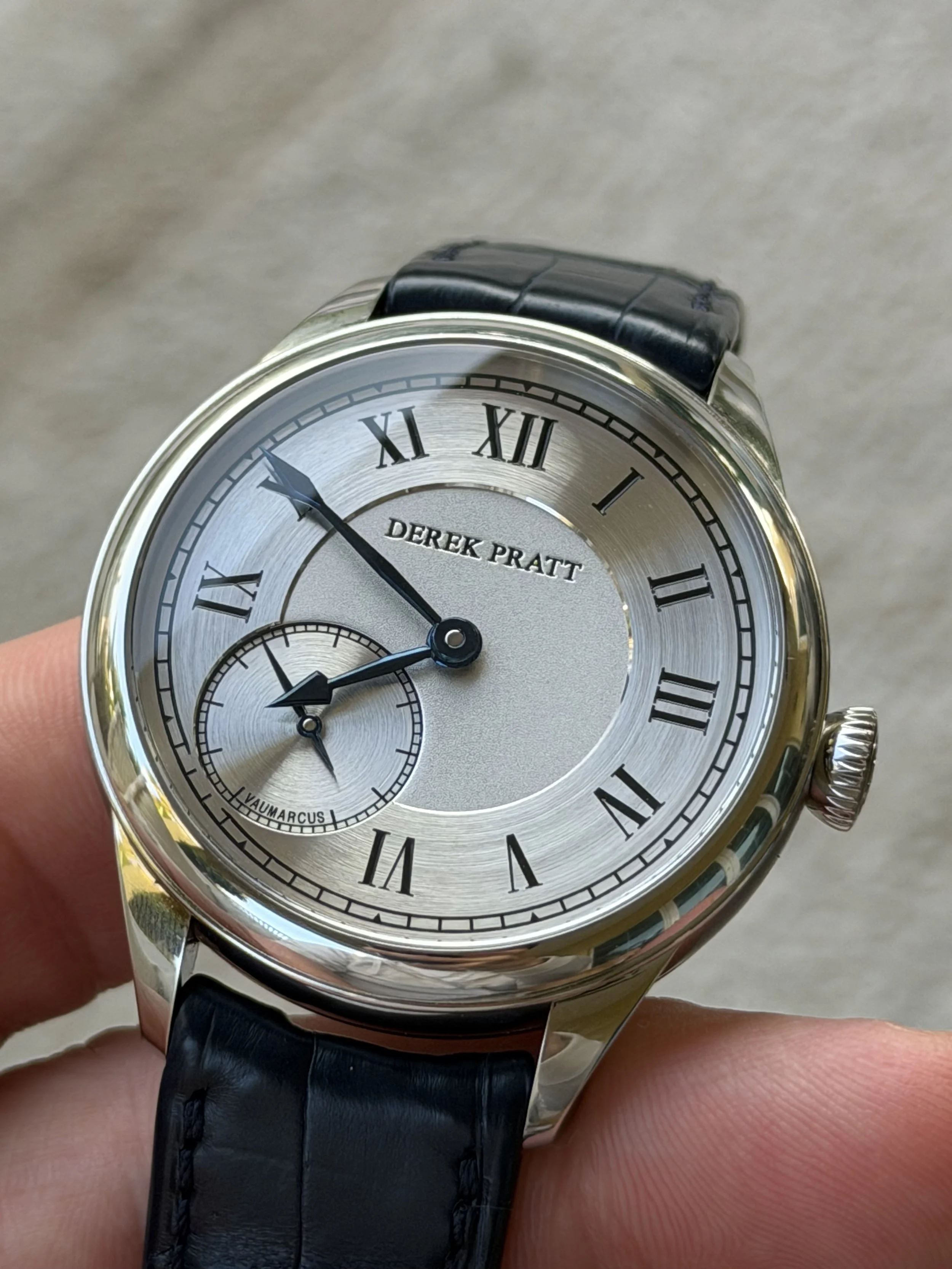 A wristwatch with a silver dial, black Roman numerals, and blue hands, featuring a subdial at 9 o'clock. The watch has a black leather strap and the brand name "Derek Pratt" on the dial.