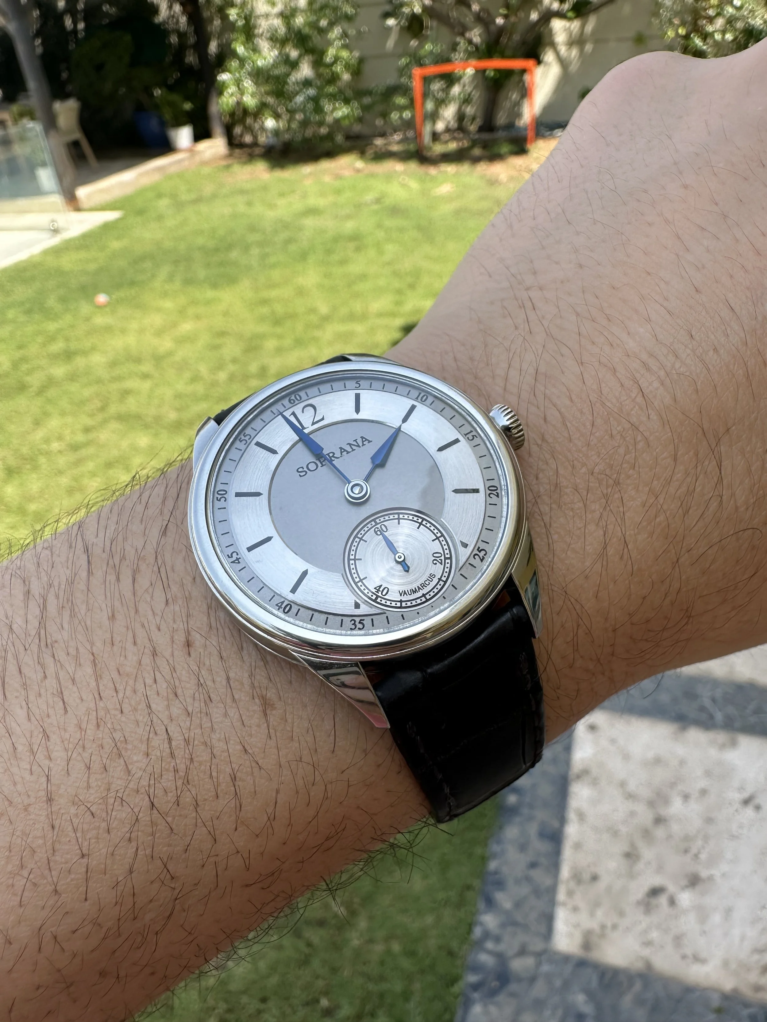 A person's wrist wearing a silver watch with a black leather strap, showing the time as 1:59. The watch face has a secondary dial indicating seconds and a digital style design with the brand name 'Sorrana.' The background is a green garden with a sma