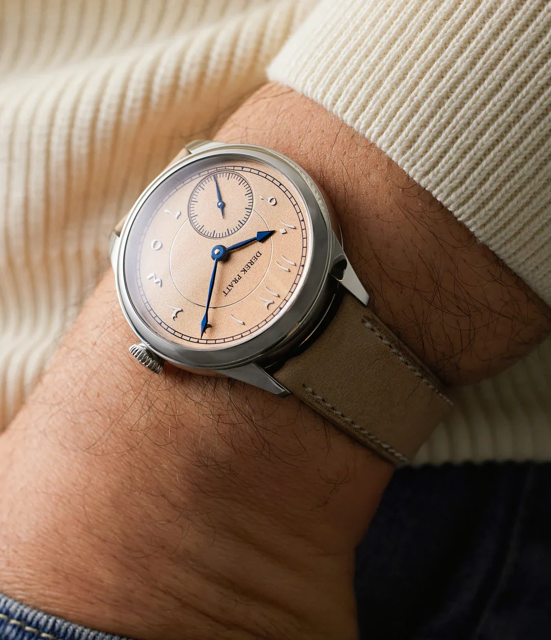 A wristwatch with a beige dial and blue hands on a person's wrist, with a beige strap and a cream-colored sweater sleeve.
