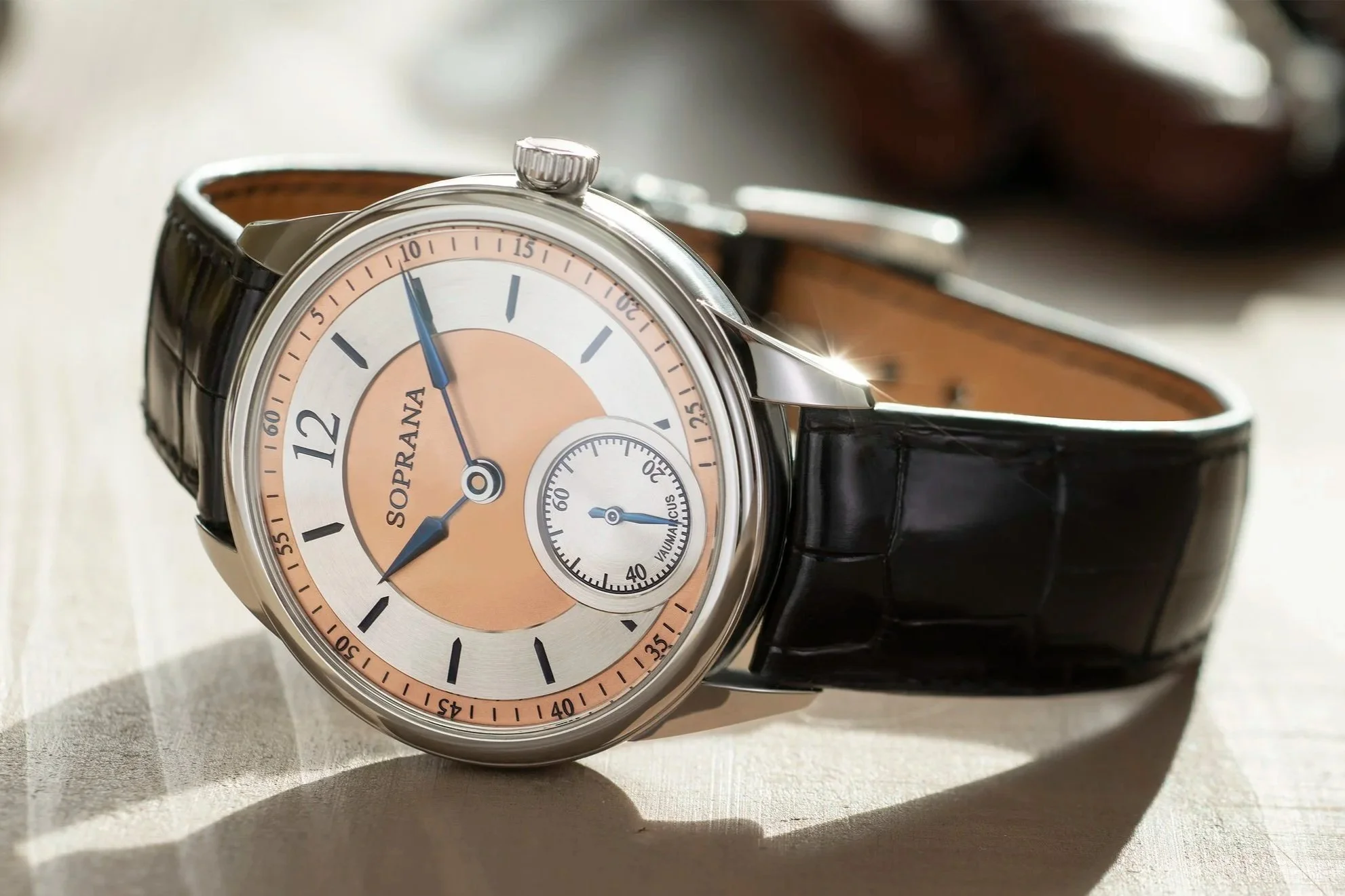 A luxury wristwatch with a round face, black leather strap, silver case, and beige and peach dial with blue hands, displaying the time and an additional small seconds sub-dial.