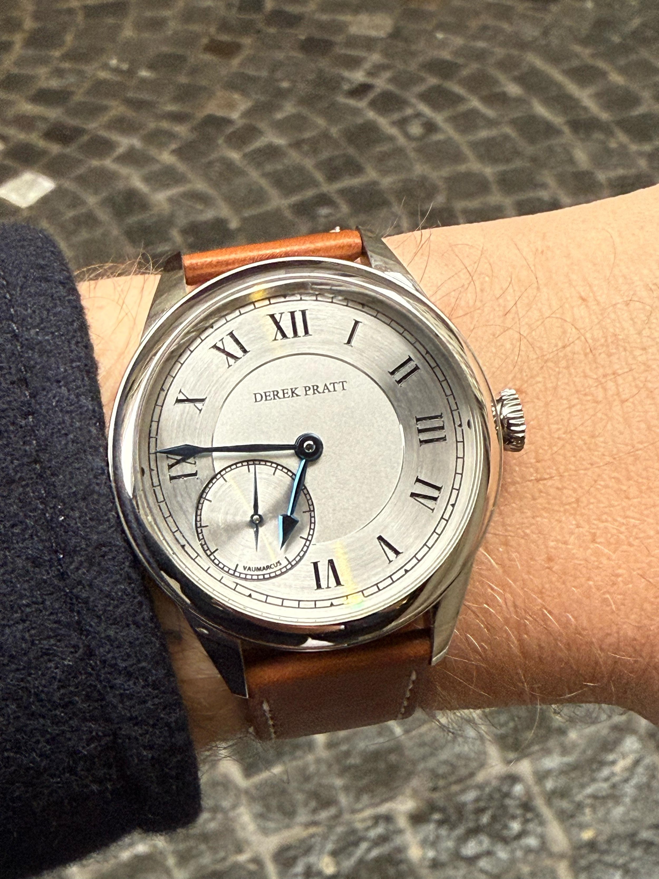 Close-up of a wristwatch on a person's wrist, showing the time as approximately 8:45, with a silver dial, Roman numeral hour markers, and a small seconds subdial at the 6 o'clock position. The watch has a brown leather strap and the person's sleeve i