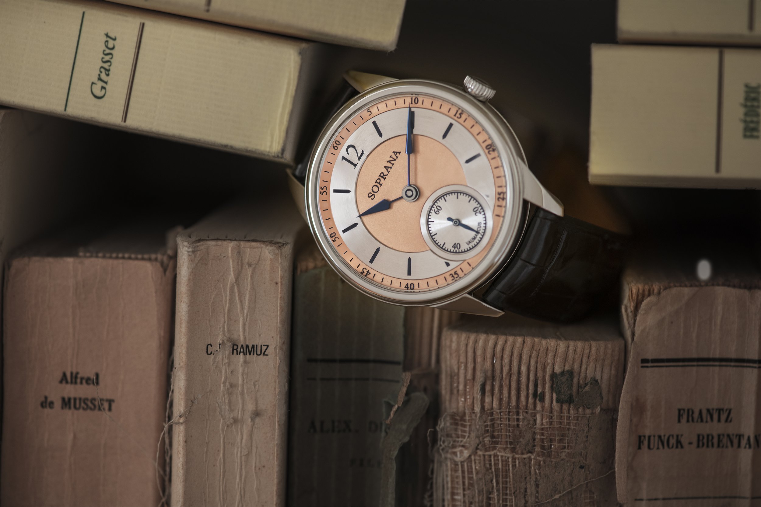 A wristwatch with a black leather strap, round face, and pink and silver dial, resting among old books on a shelf.