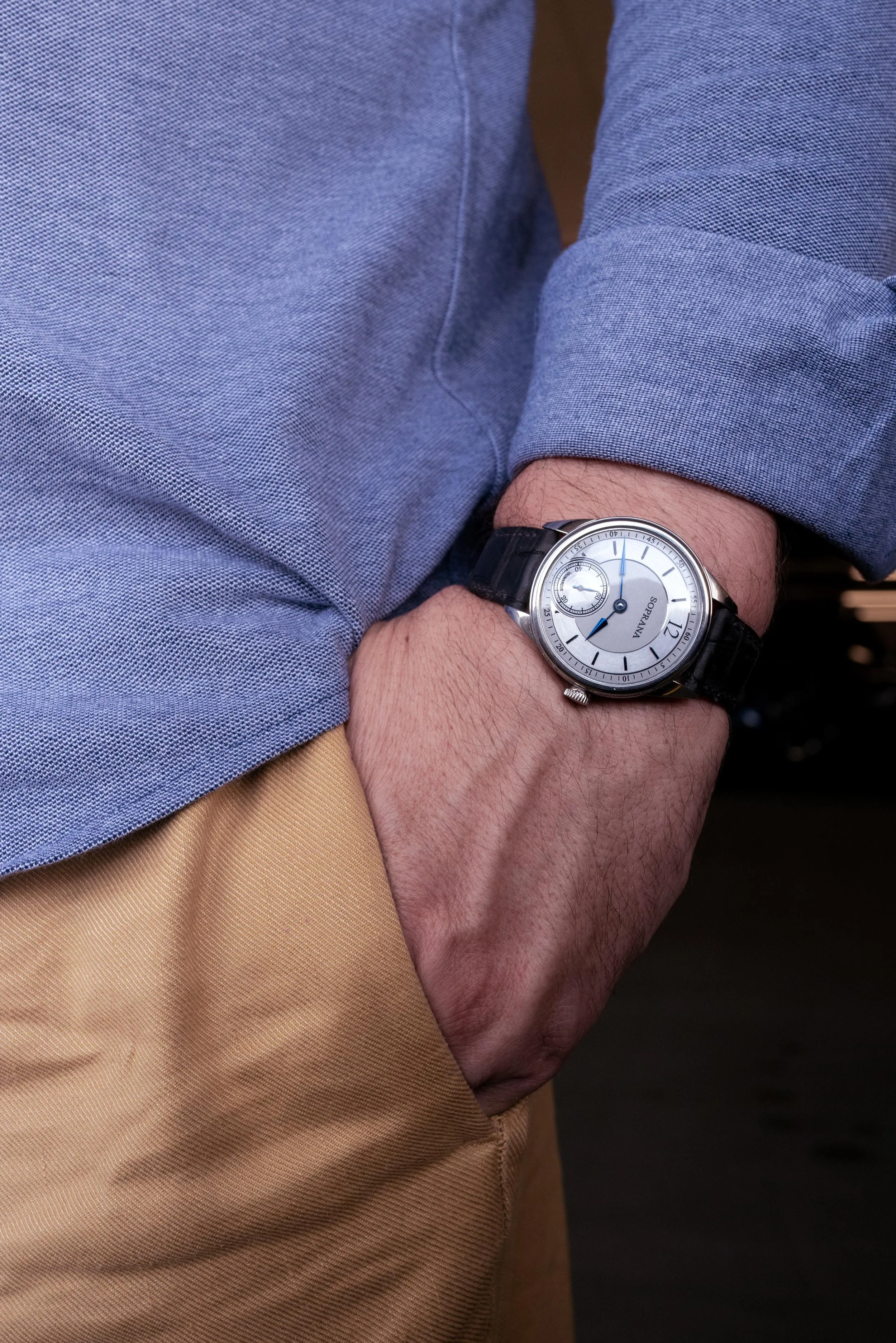 Close-up of a person's wrist wearing a silver watch with a black leather strap, a light gray dial, and a small second subdial, with the person's hand in their pocket, dressed in a blue shirt and tan pants.