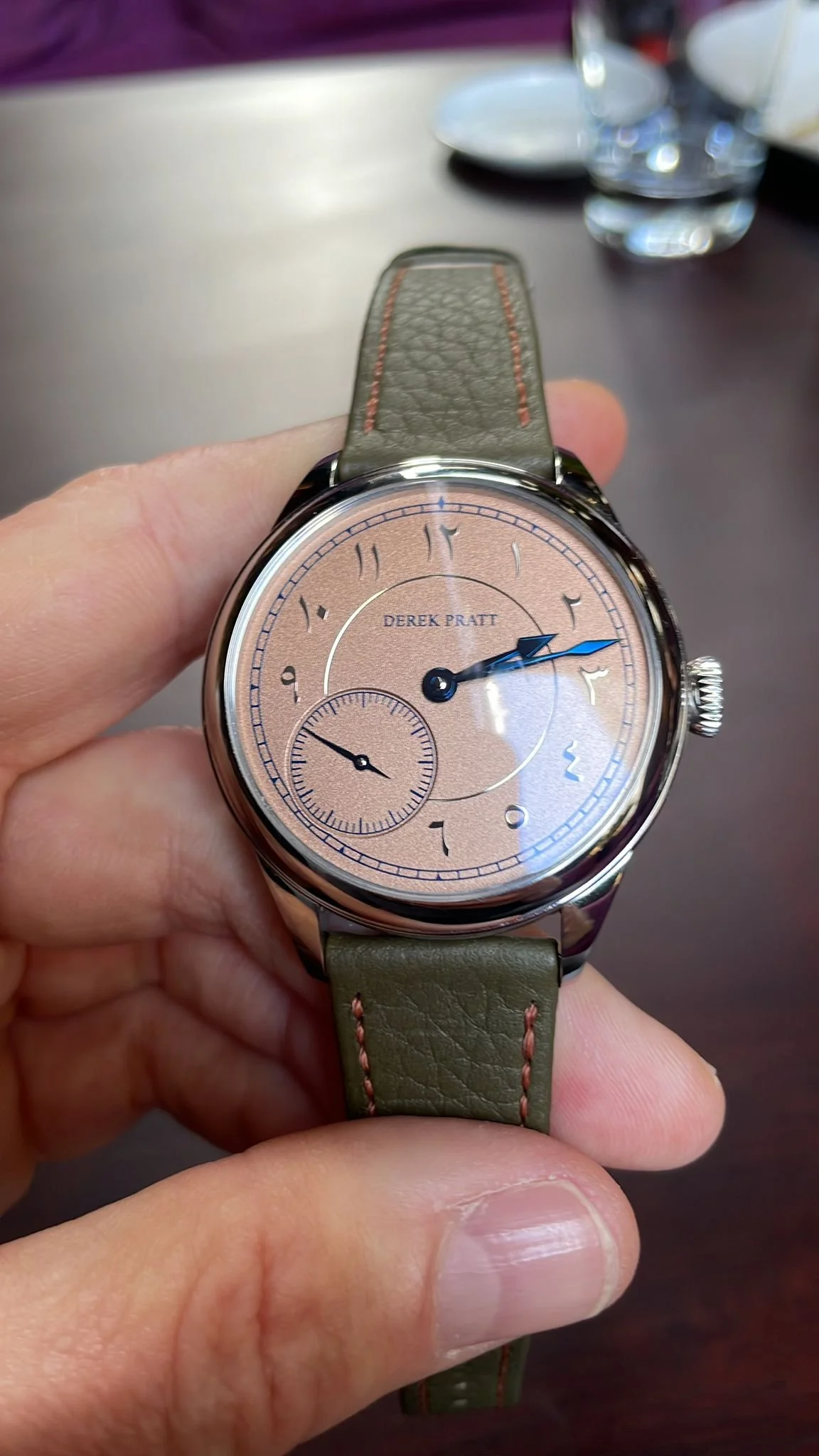 A Derek Pratt wristwatch with a beige dial, black and blue hands, and a small subdial at the six o'clock position, attached to a green leather strap with red stitching.