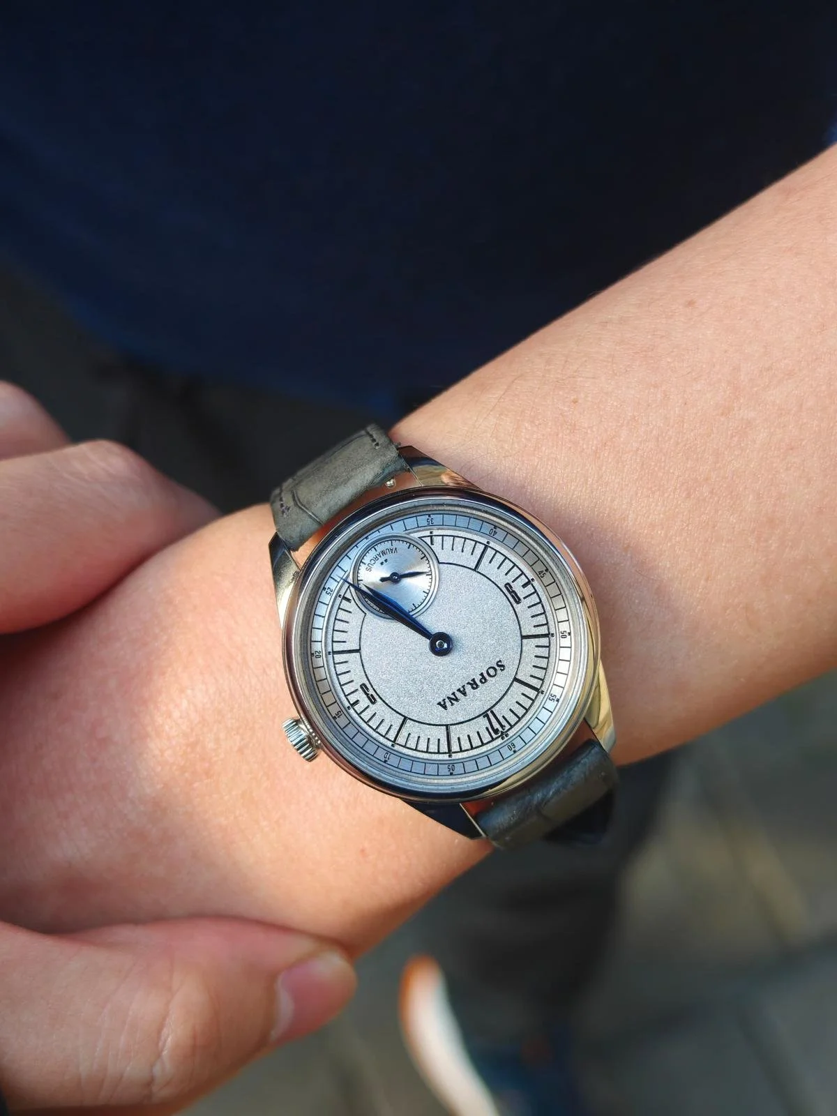 A person's wrist wearing a silver watch with a black leather strap, showing the time as 8:45, with a small subdial for seconds.