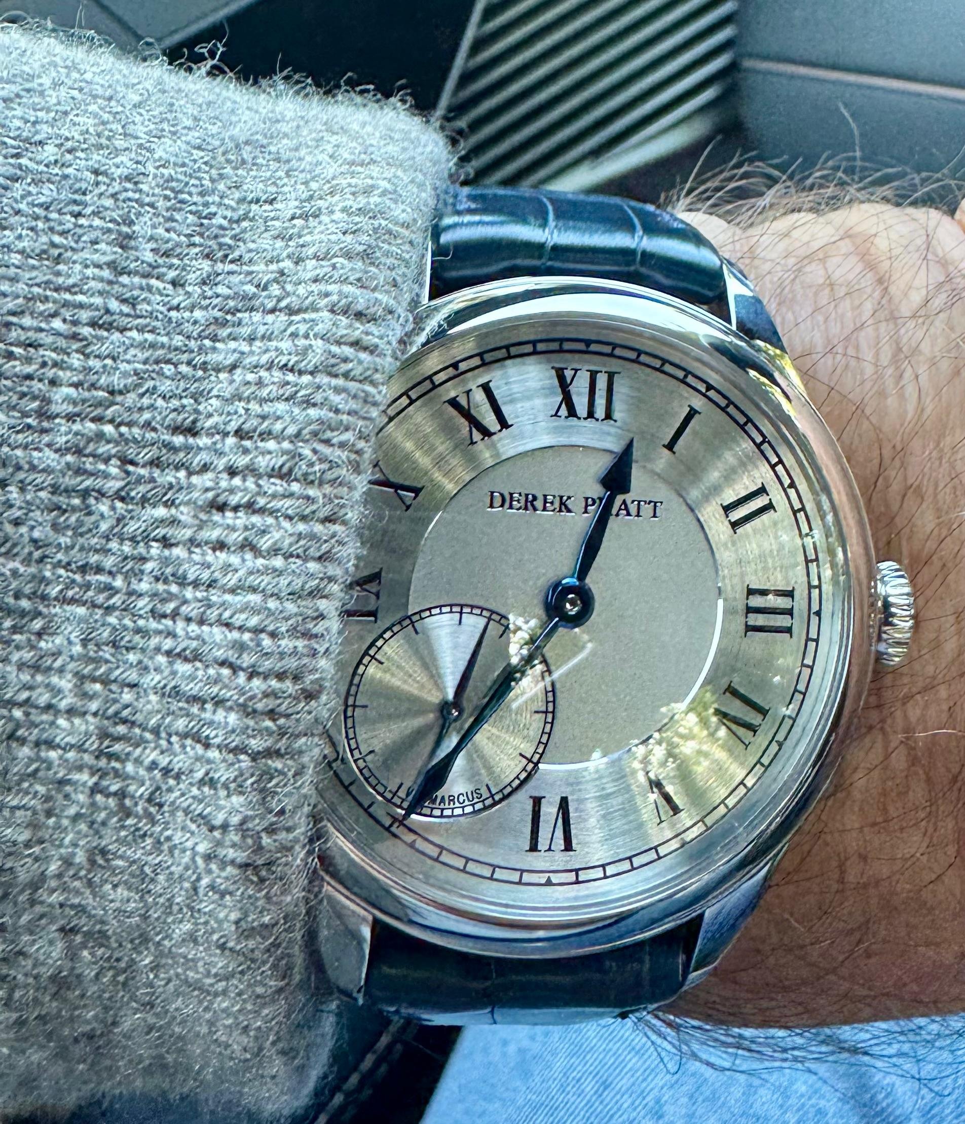 A close-up of a wristwatch with a silver dial, Roman numerals, black hands, and a black leather strap, worn on a person's wrist.