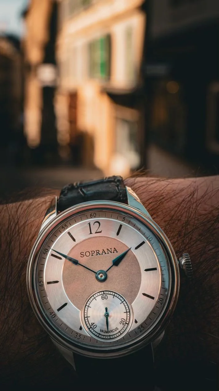 A wristwatch with the brand name SOPRANA, showing the time as approximately 1:50, with a background of bookshelves in a possibly cozy indoor setting.