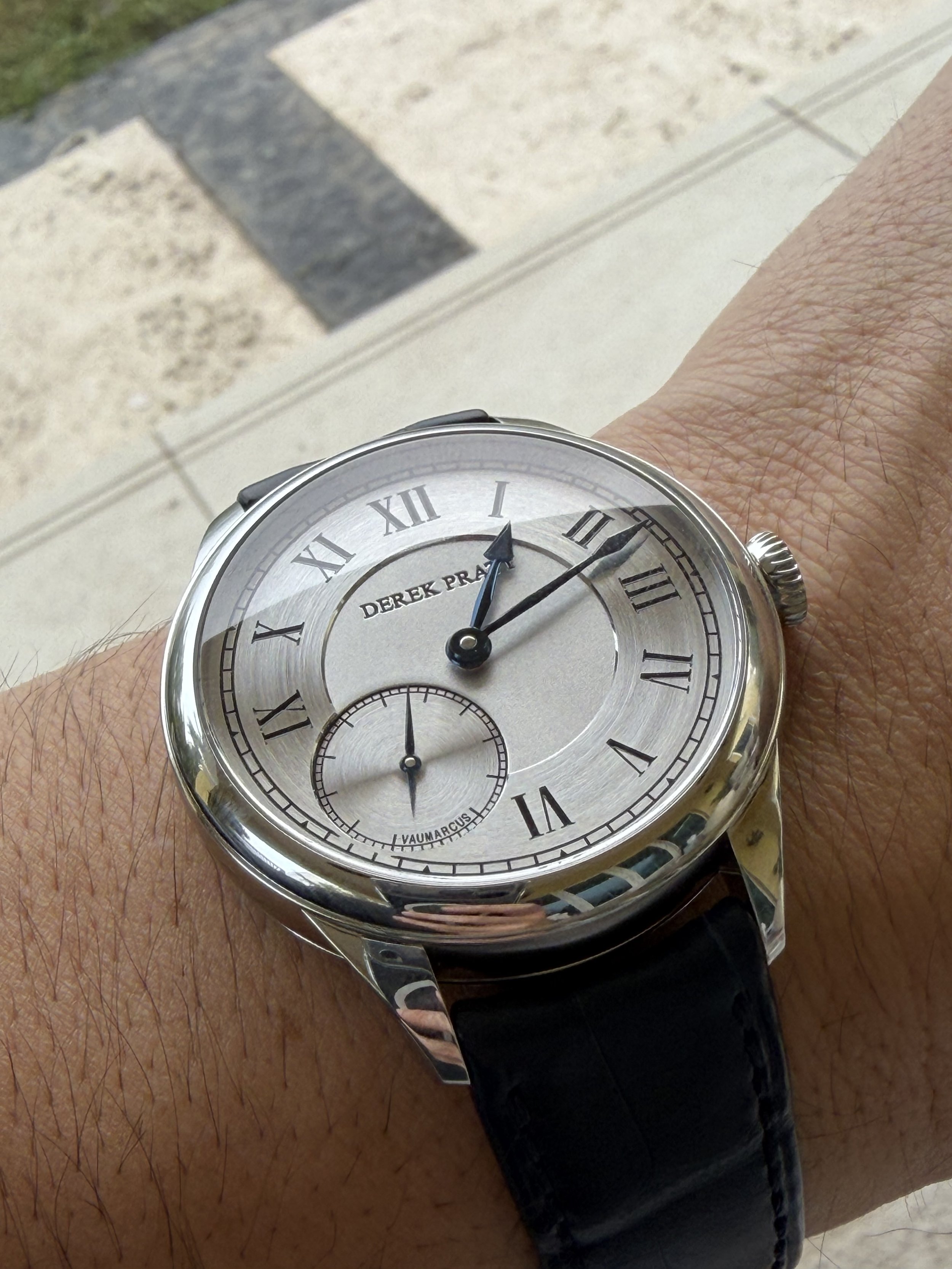 A wristwatch with a silver face, black Roman numeral hour markers, and black minute and second hands. It has a small seconds subdial at the six o'clock position and the brand name 'DEREK PRATT' on the face. The watch is worn on a person's wrist with 