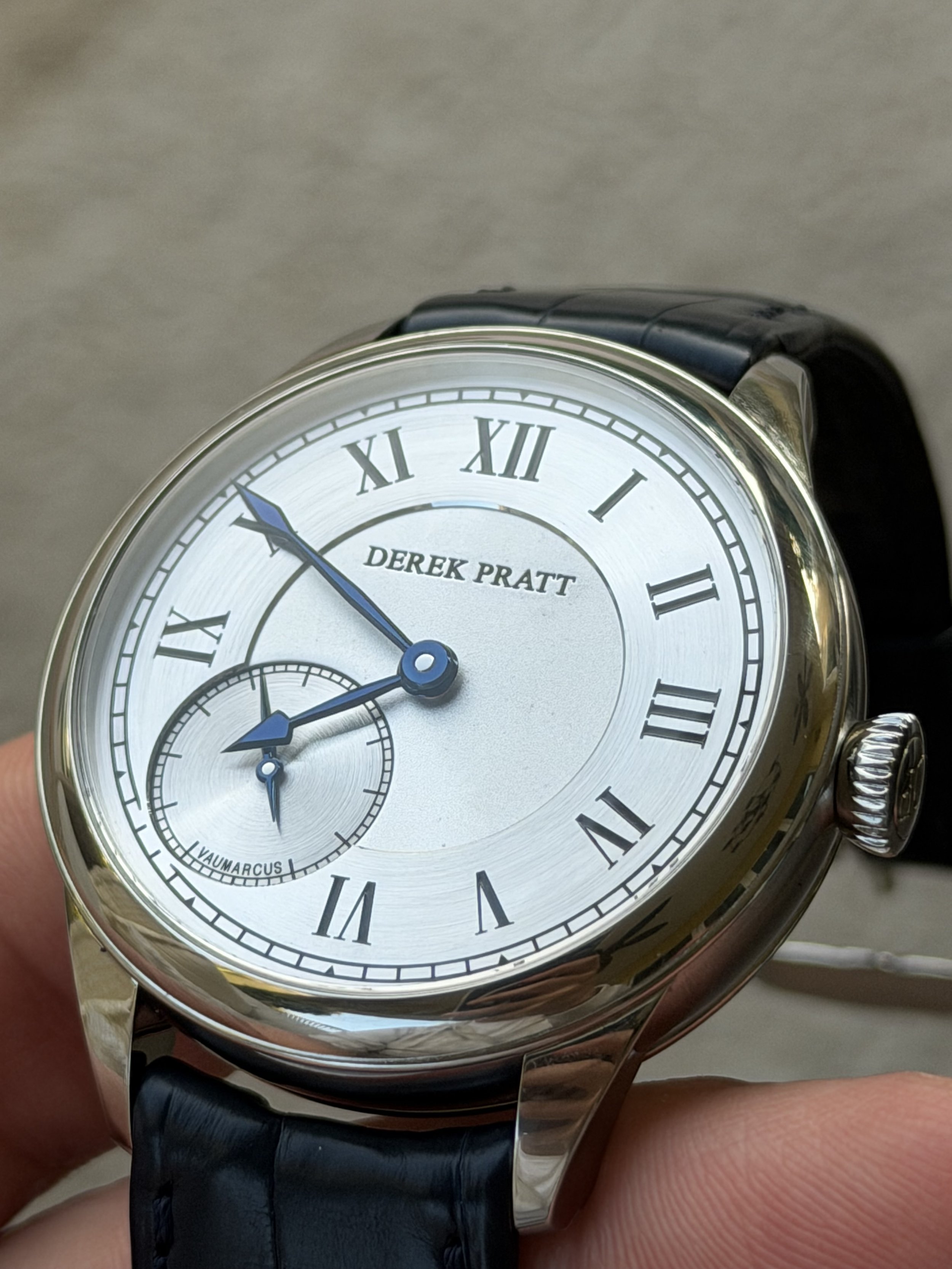 A wristwatch with a white dial, Roman numeral hour markers, and blue hands, showing the time as 9:47, with a subdial at 8 o'clock, on a black leather strap. The name 'DEREK PRATT' is printed on the dial.