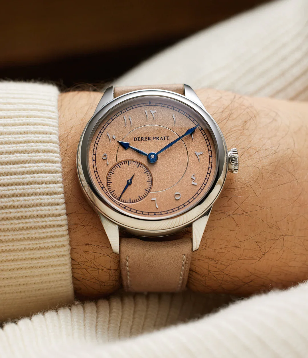 A wristwatch with a beige face and blue hands, showing the time as 10:10, on a beige strap, worn on a person's wrist, with the brand name 'Derek Pratt' on the dial.