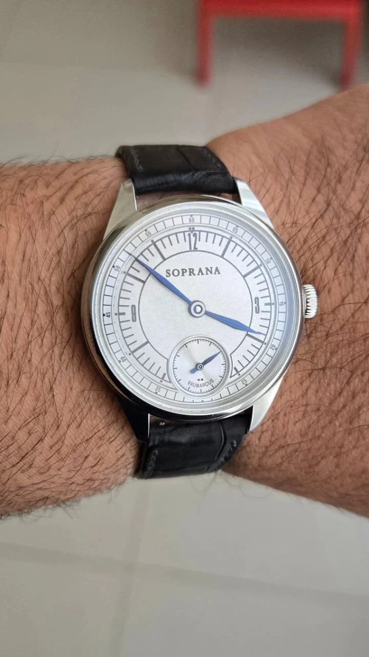 A wristwatch with a silver case, black leather strap, and a white dial showing the brand Soprana. The watch has blue hour and minute hands and a small seconds subdial at the bottom, labeled VauMarcus.