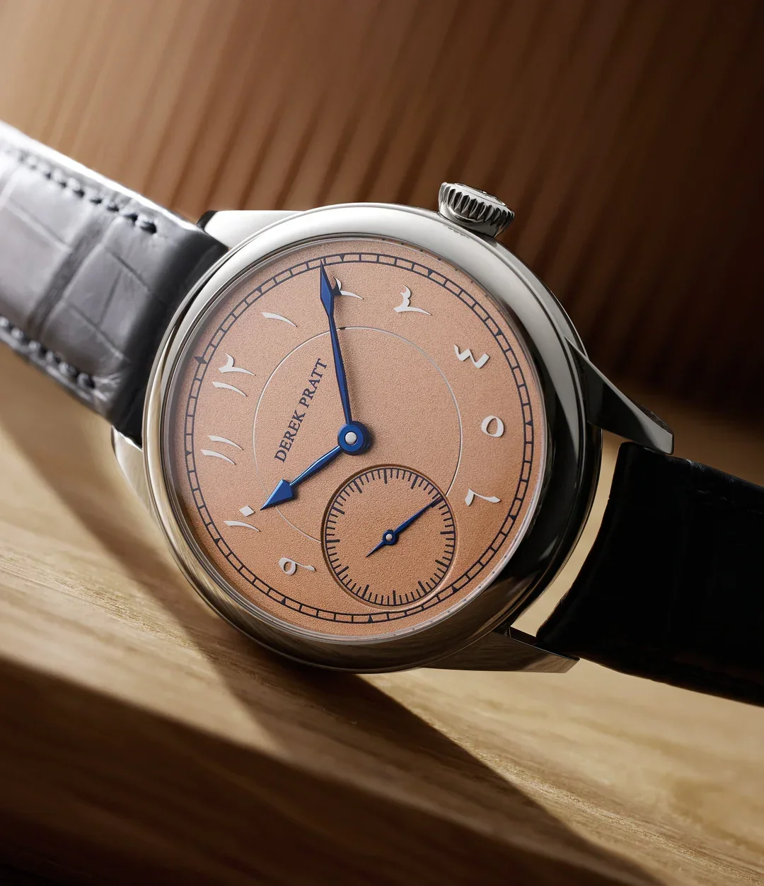 Close-up of a luxury wristwatch with a beige dial, blue hands, and black leather strap. The watch face shows the brand name 'DEREK PRATT' and features a small subdial at the 6 o'clock position.