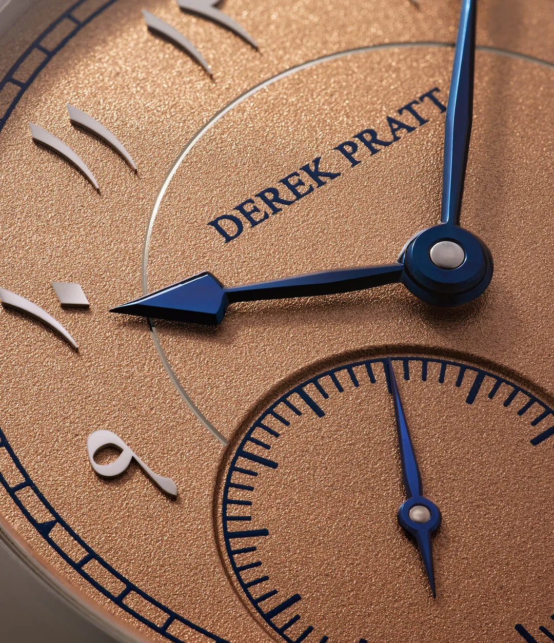 Close-up of a luxury watch face with gold textured background, blue hands, and white markers, displaying the brand name Derek Pratt.